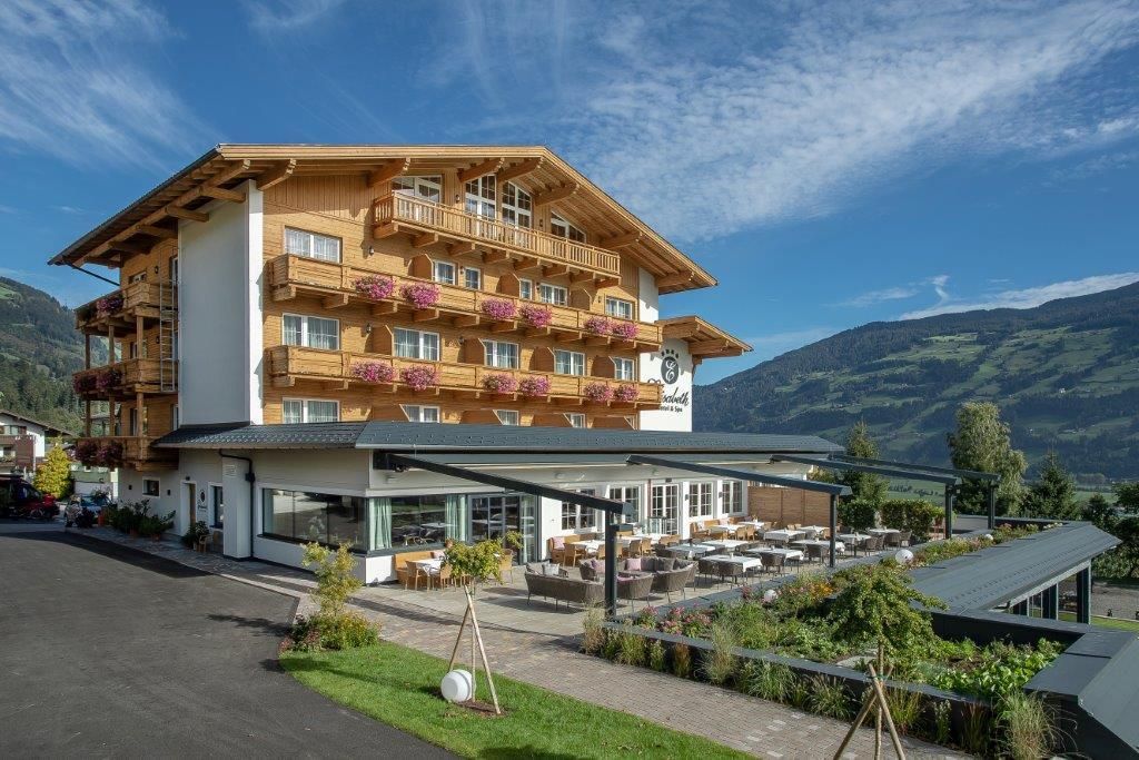 A modern hotel with wooden paneling and balconies decorated with flowers. In the background, green hills and a blue sky stretch out.