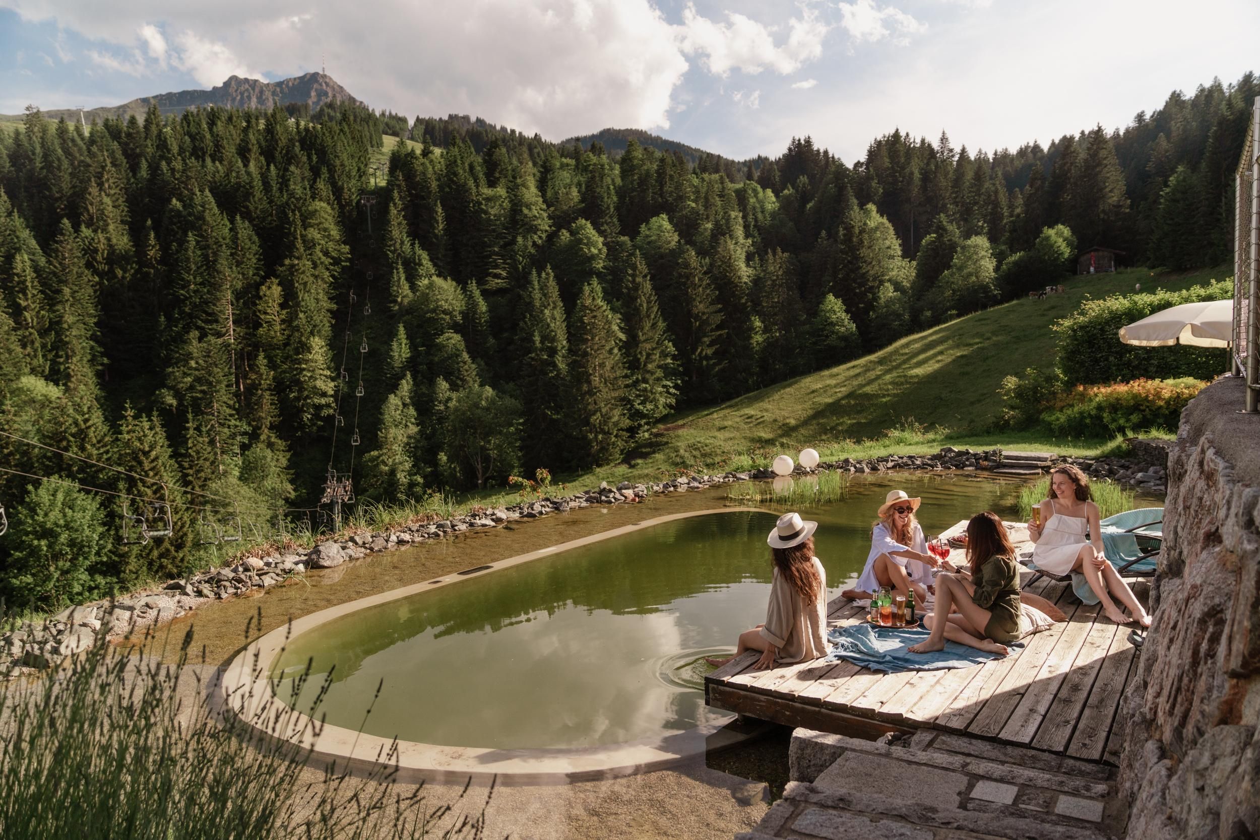 A group of women relaxes at the edge of a pond in a picturesque natural landscape. Surrounded by green forests and mountains, they enjoy their time in the sun.