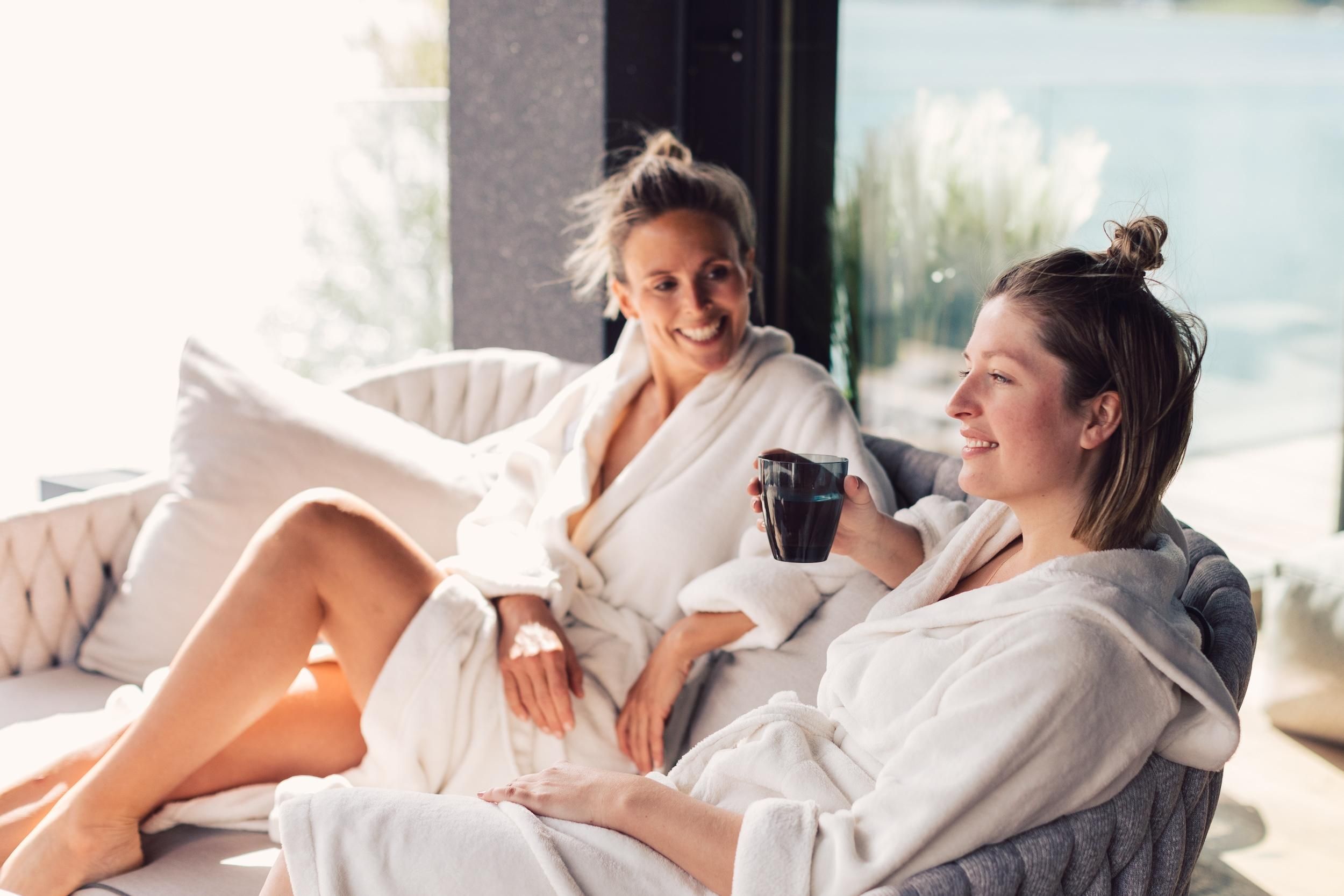 Two women in white bathrobes relax on a cozy couch. One woman is holding a drink in her hand and smiling.
