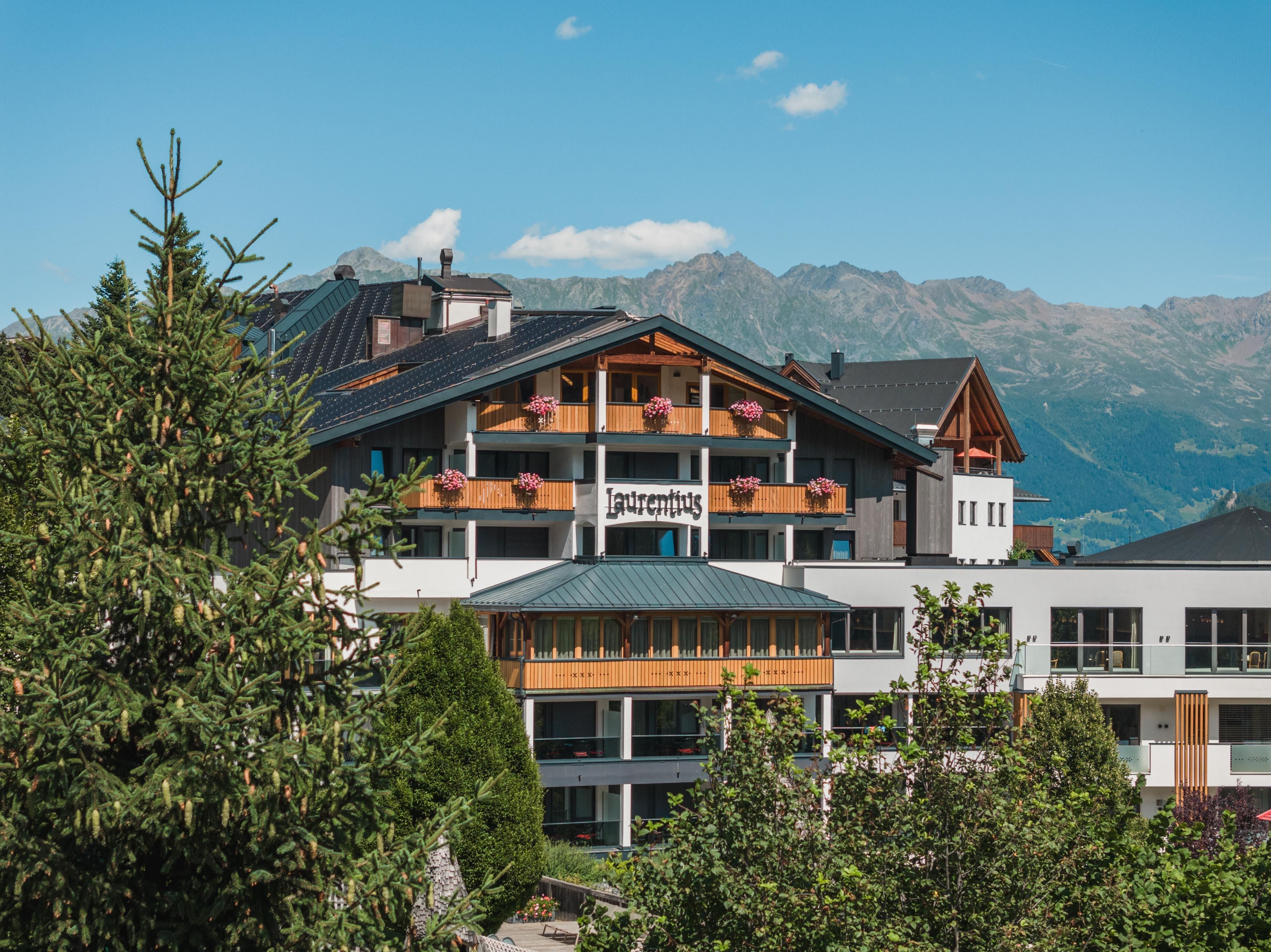 A cozy hotel in the mountains, surrounded by green trees. In the background, majestic mountains and a clear sky can be seen.
