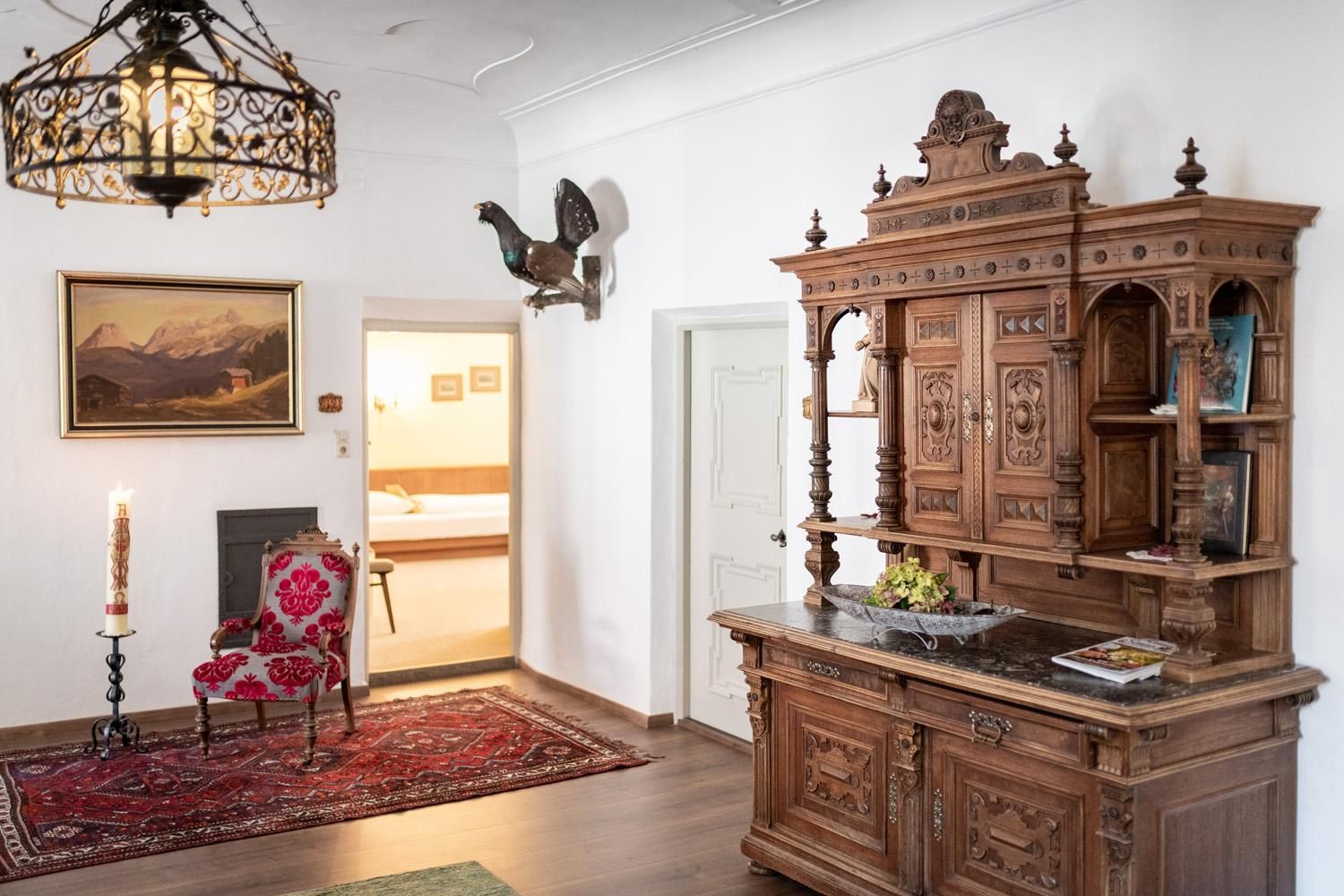 A stylish room with an antique wooden cupboard and an elegant armchair. A painting and a decorative figure hang on the wall.