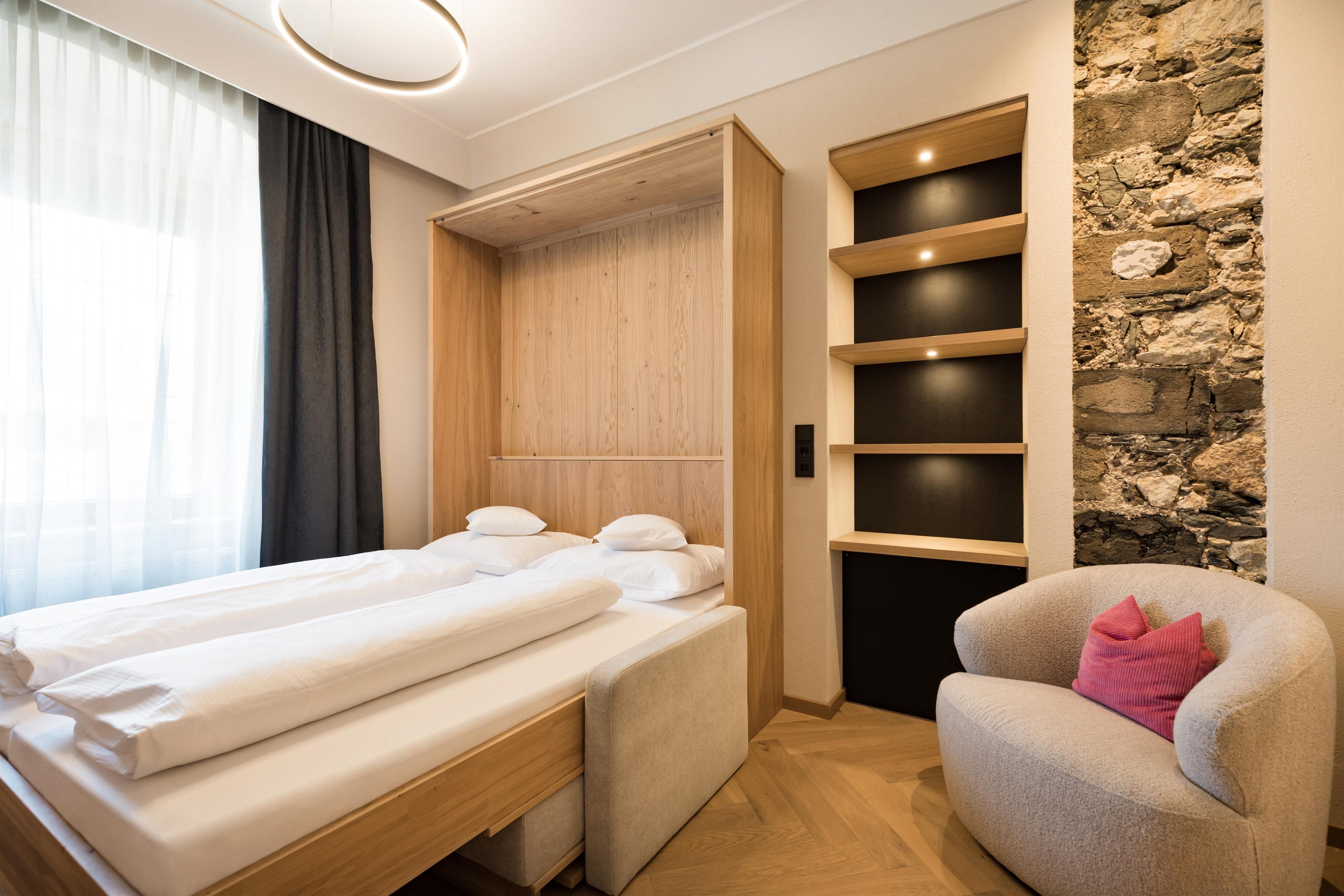 A modern bedroom with a cozy bed and a chair. The walls feature a mix of wood and stone, with plenty of natural light coming through the window.