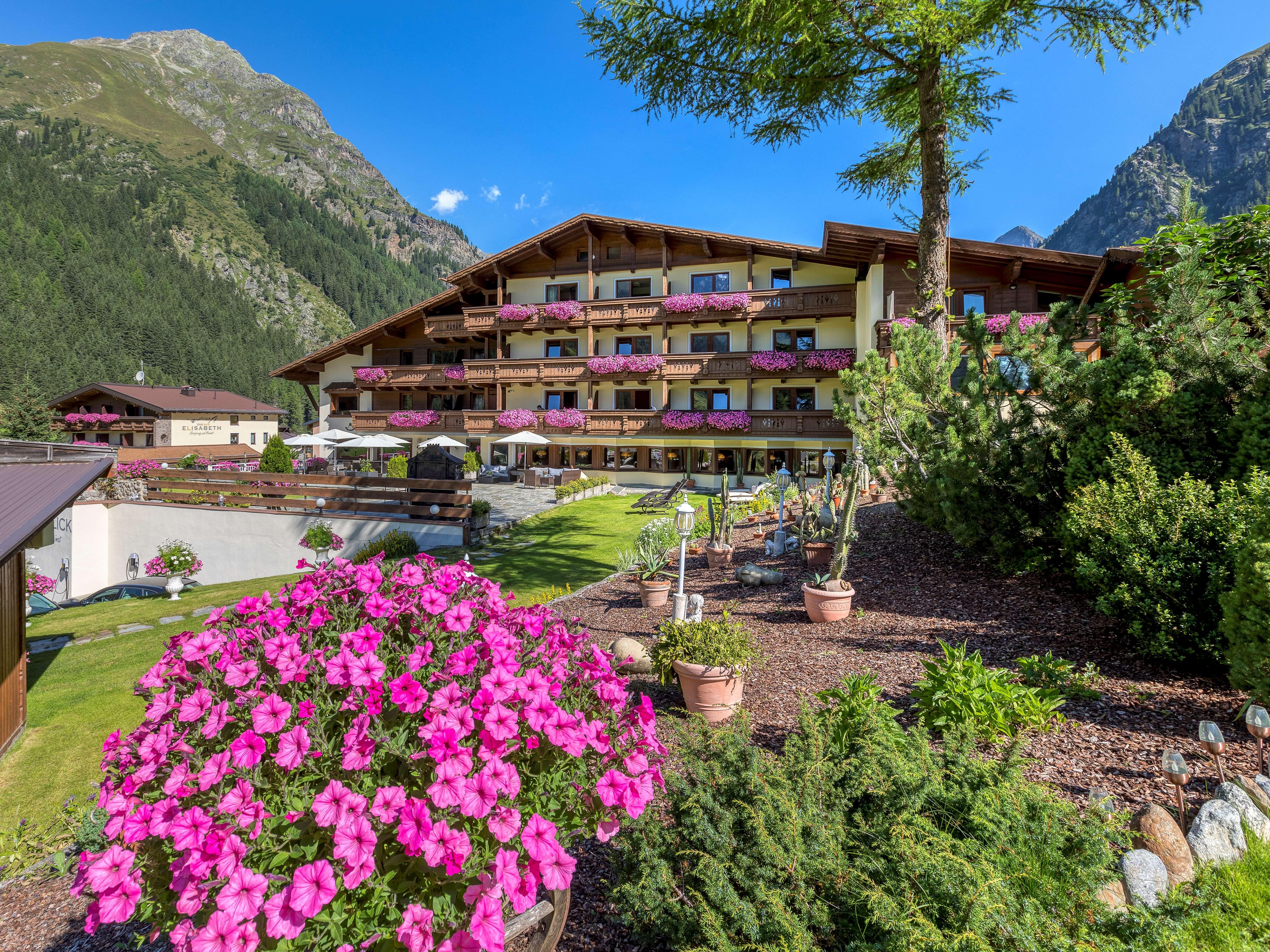 A hotel building surrounded by green mountains and colorful flowers. Pink plants are blooming on the balconies and the surroundings are inviting.