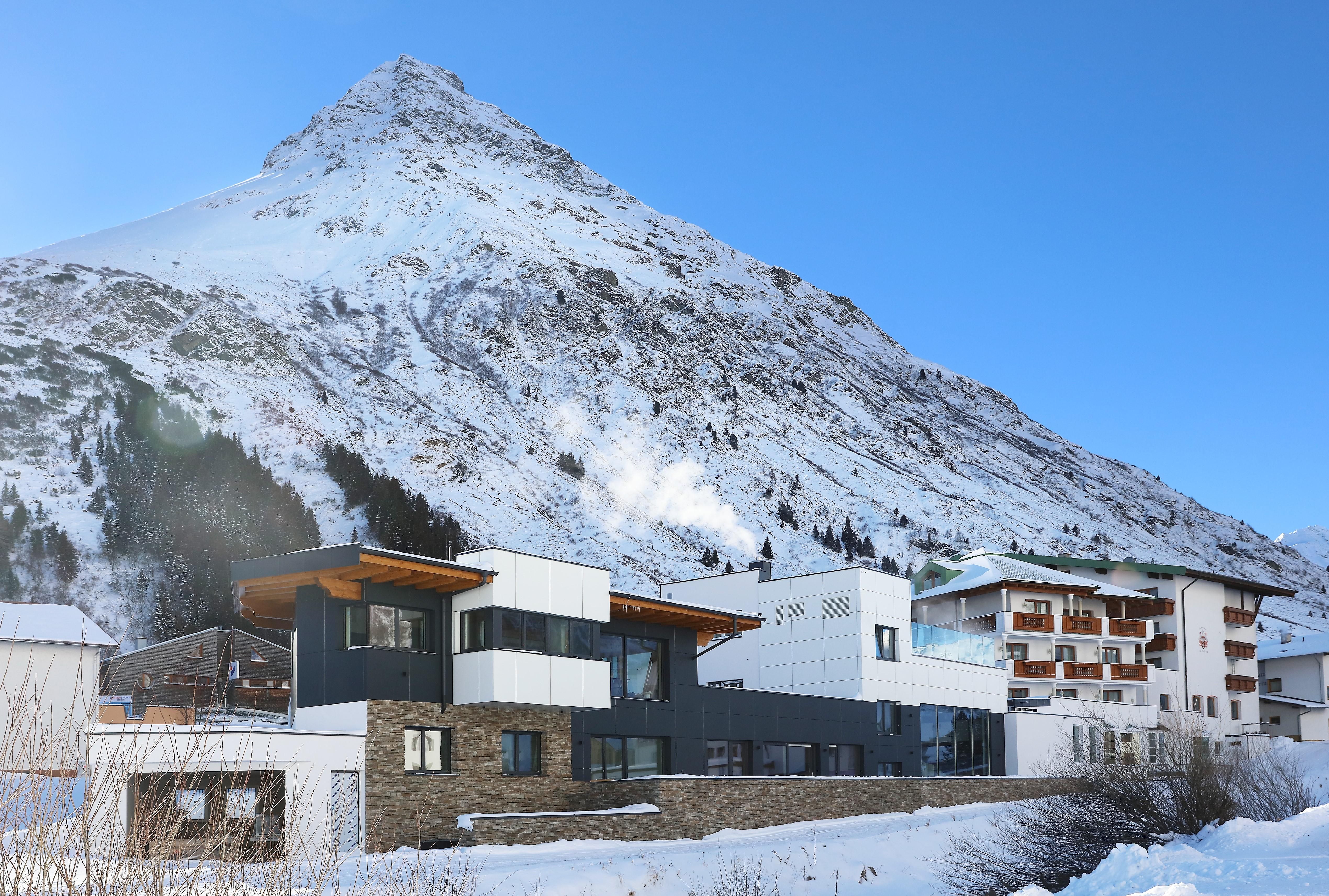 A modern building in the foreground, surrounded by snow-covered mountains. The clear blue sky completes the winter landscape.