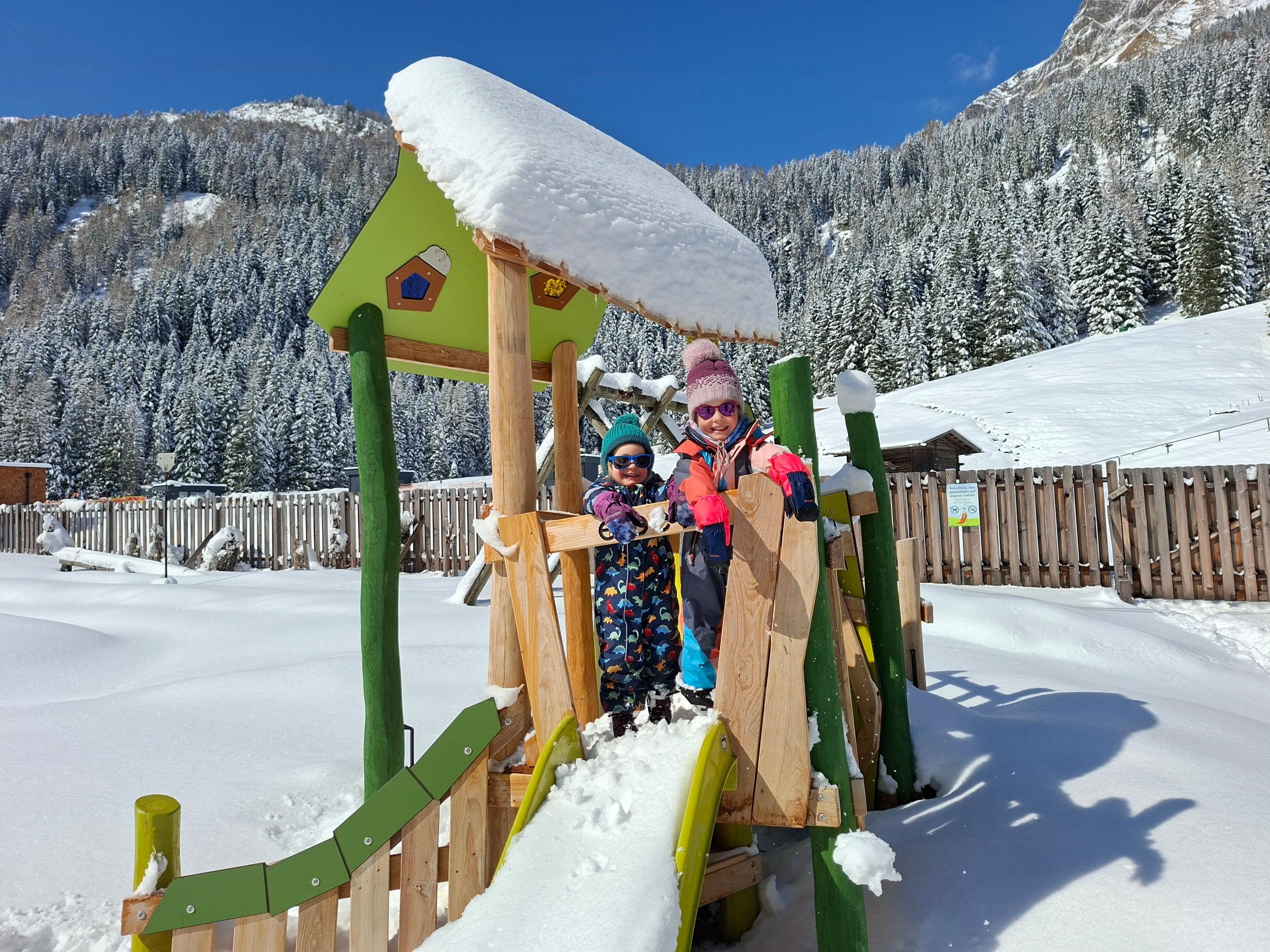 Two children are playing on a playground in the snow. In the background, there are snow-covered mountains and a blue sky.