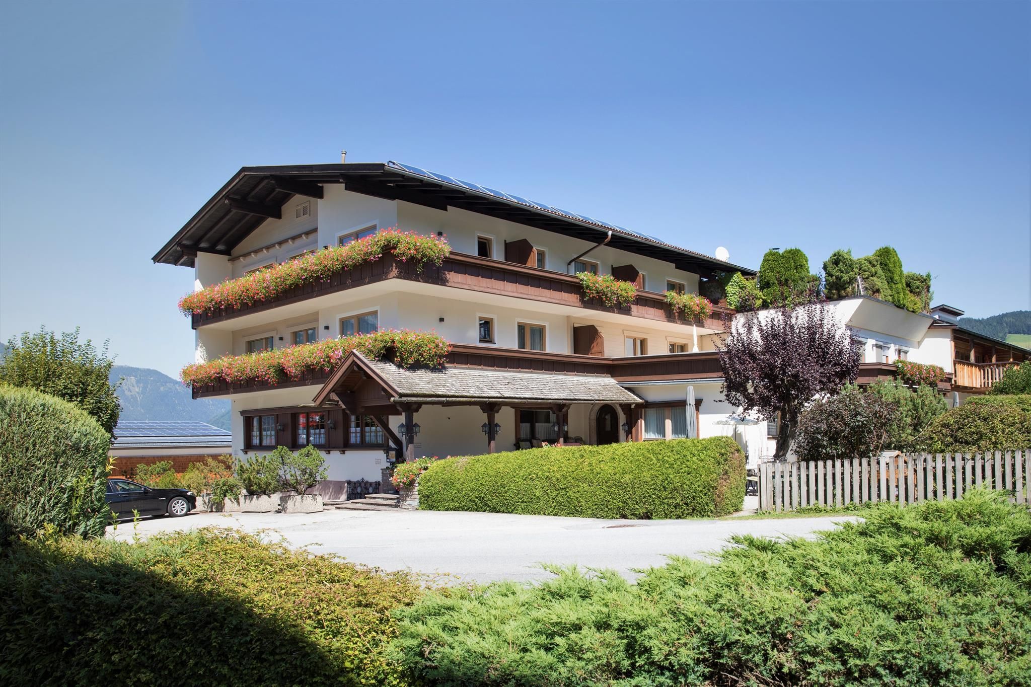 A modern building with several floors, surrounded by a well-kept garden. The facade is adorned with plants, and there is a clear blue sky in the background.
