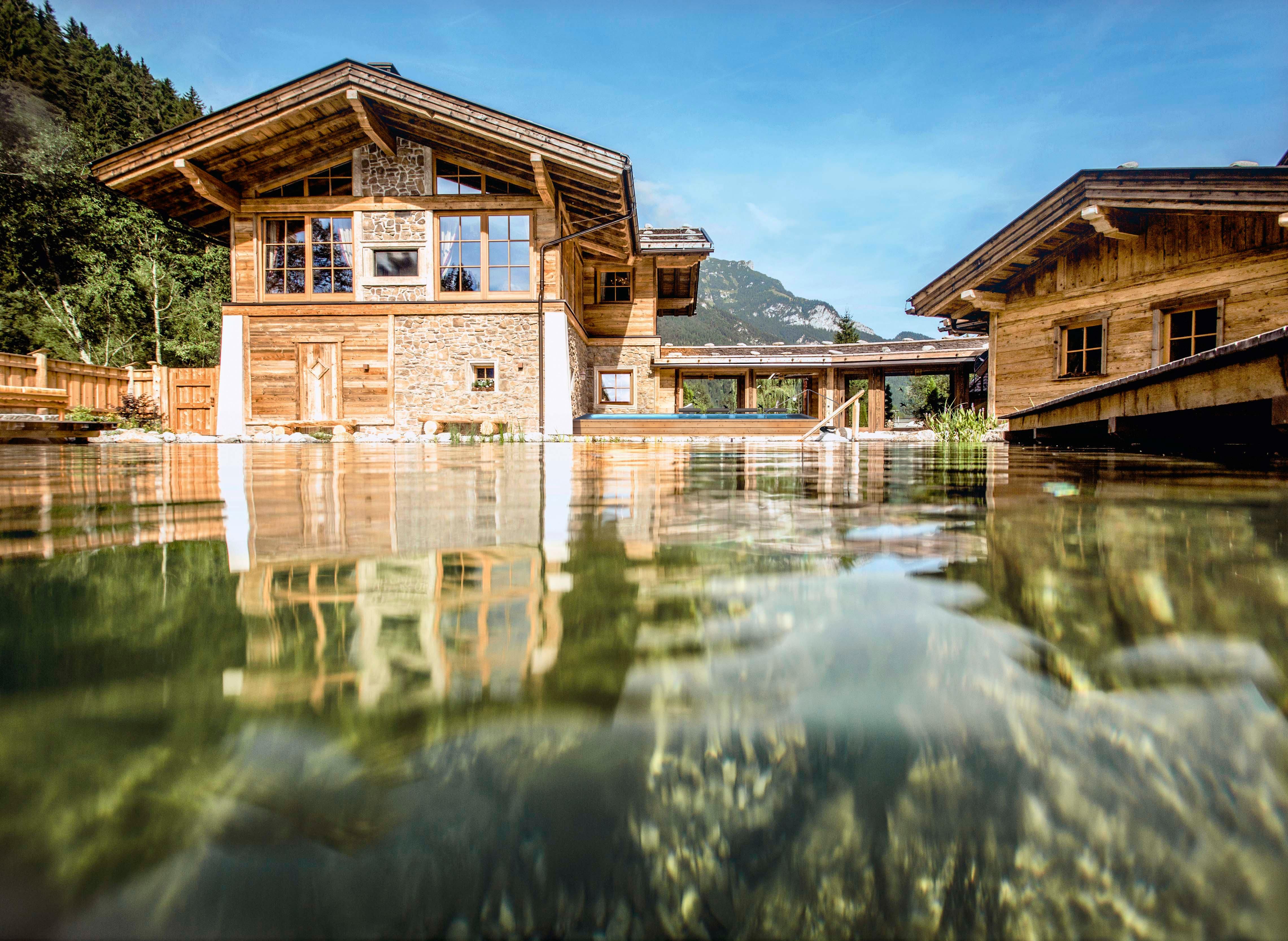 A modern wooden house by the water with large windows and a beautiful surroundings. In the background, forested mountains can be seen.