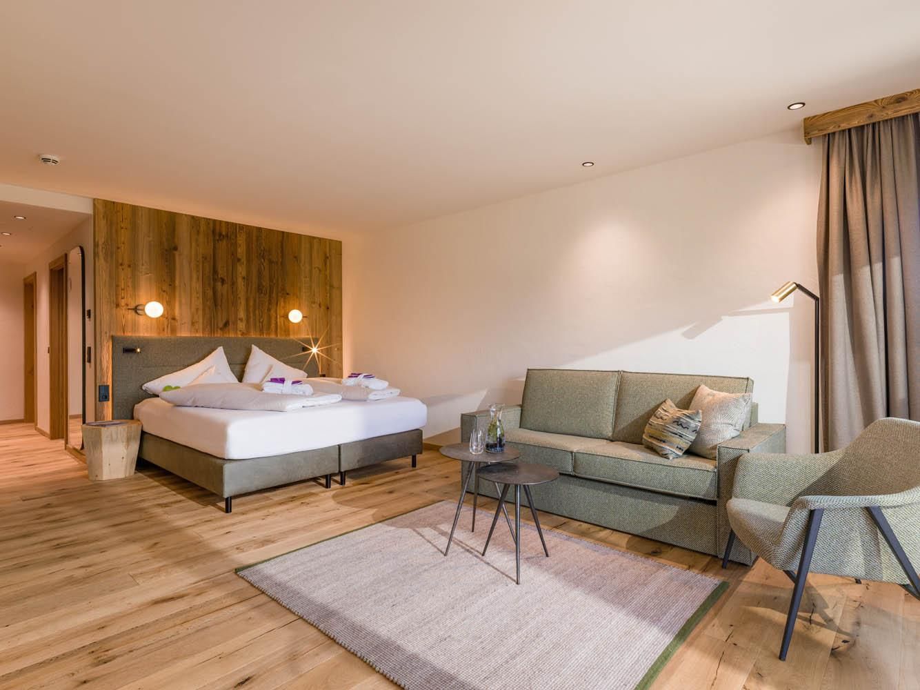 A modern hotel room with a comfortable bed and a cozy seating area. The room features wood and gray tones that create a relaxed atmosphere.