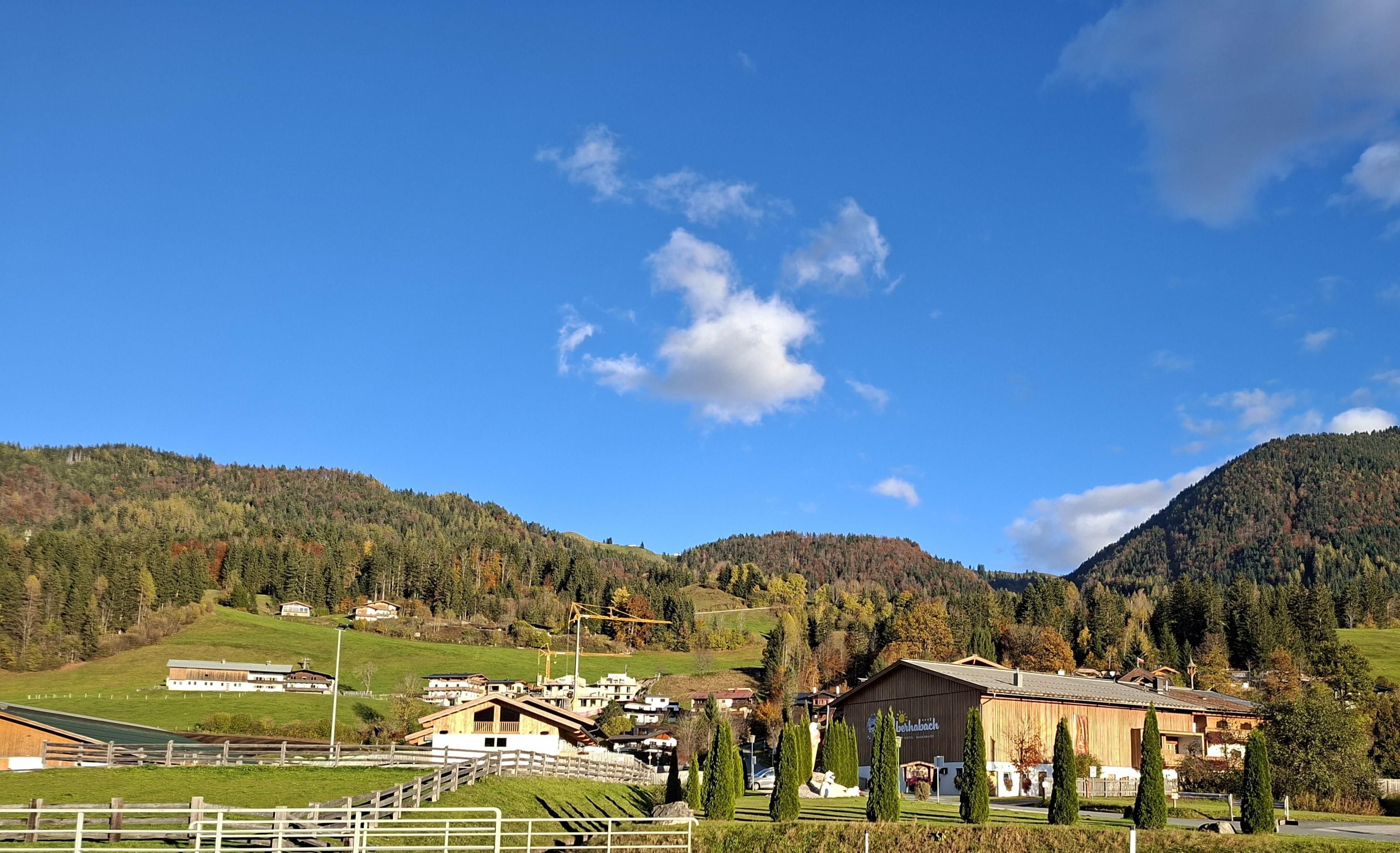 A picturesque landscape with gentle hills and a clear blue sky. In the foreground, there are cozy houses and well-maintained trees.
