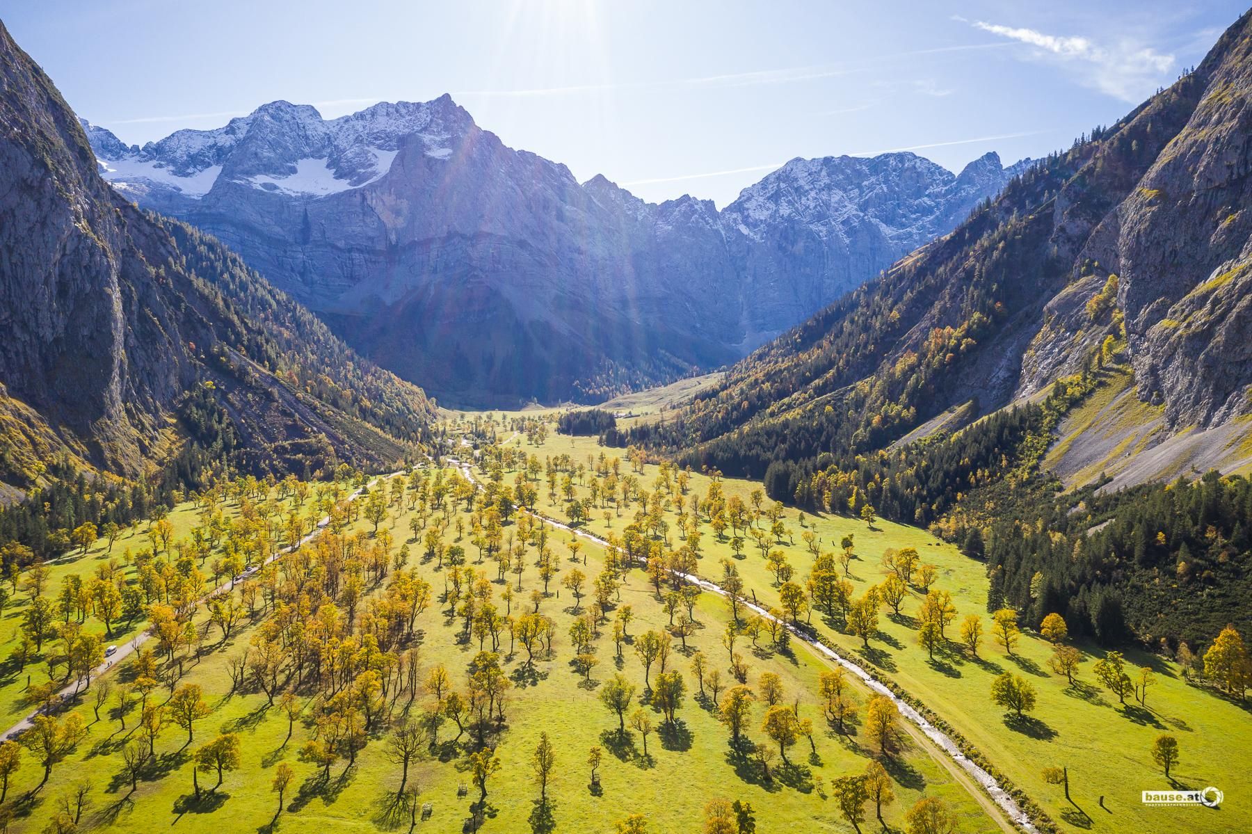 A breathtaking mountain landscape with green meadows and autumn-colored trees. In the background, majestic mountains rise under a clear blue sky.