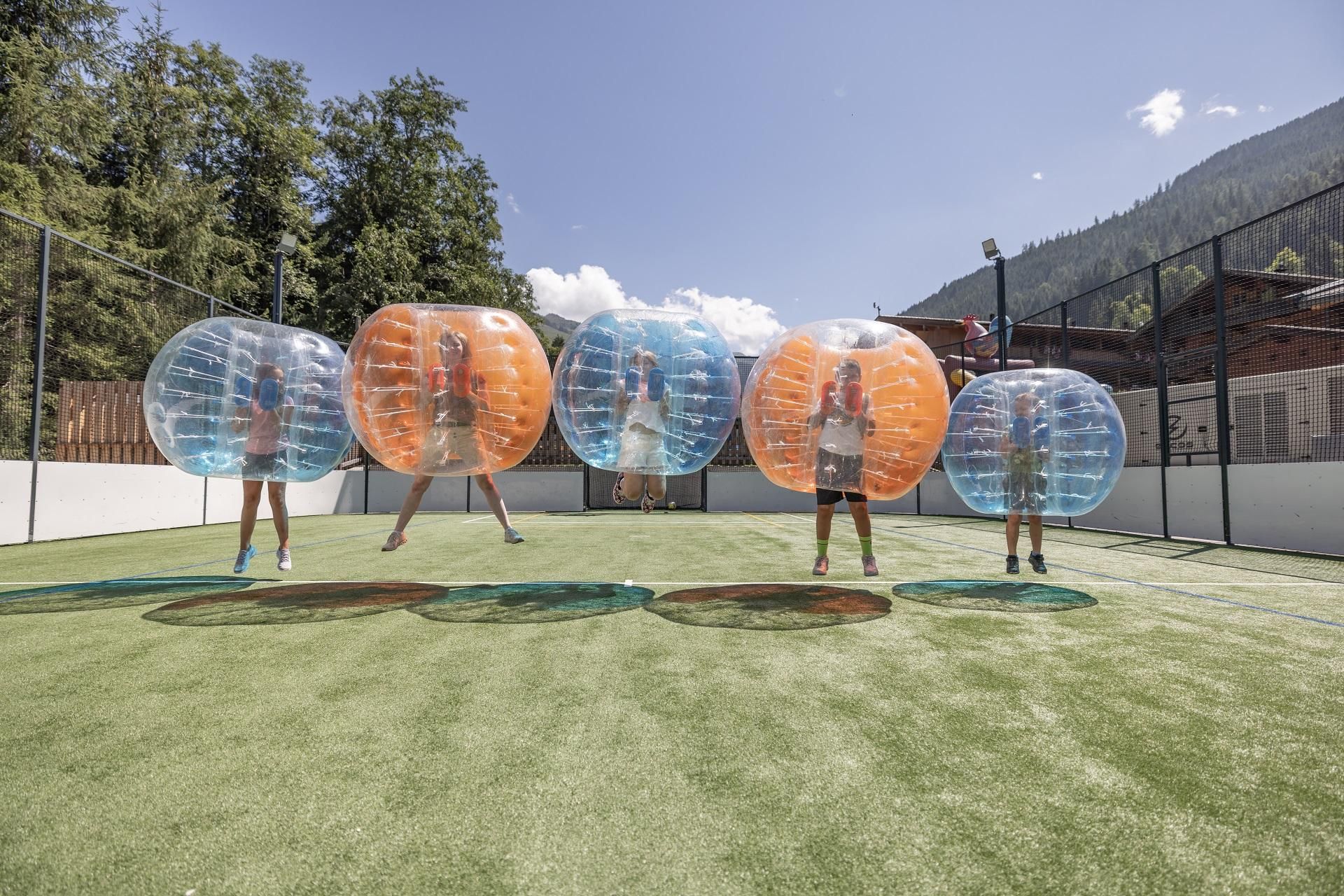 Four people are standing in colorful balloons on a sports field. The sky is clear and the surroundings are green.