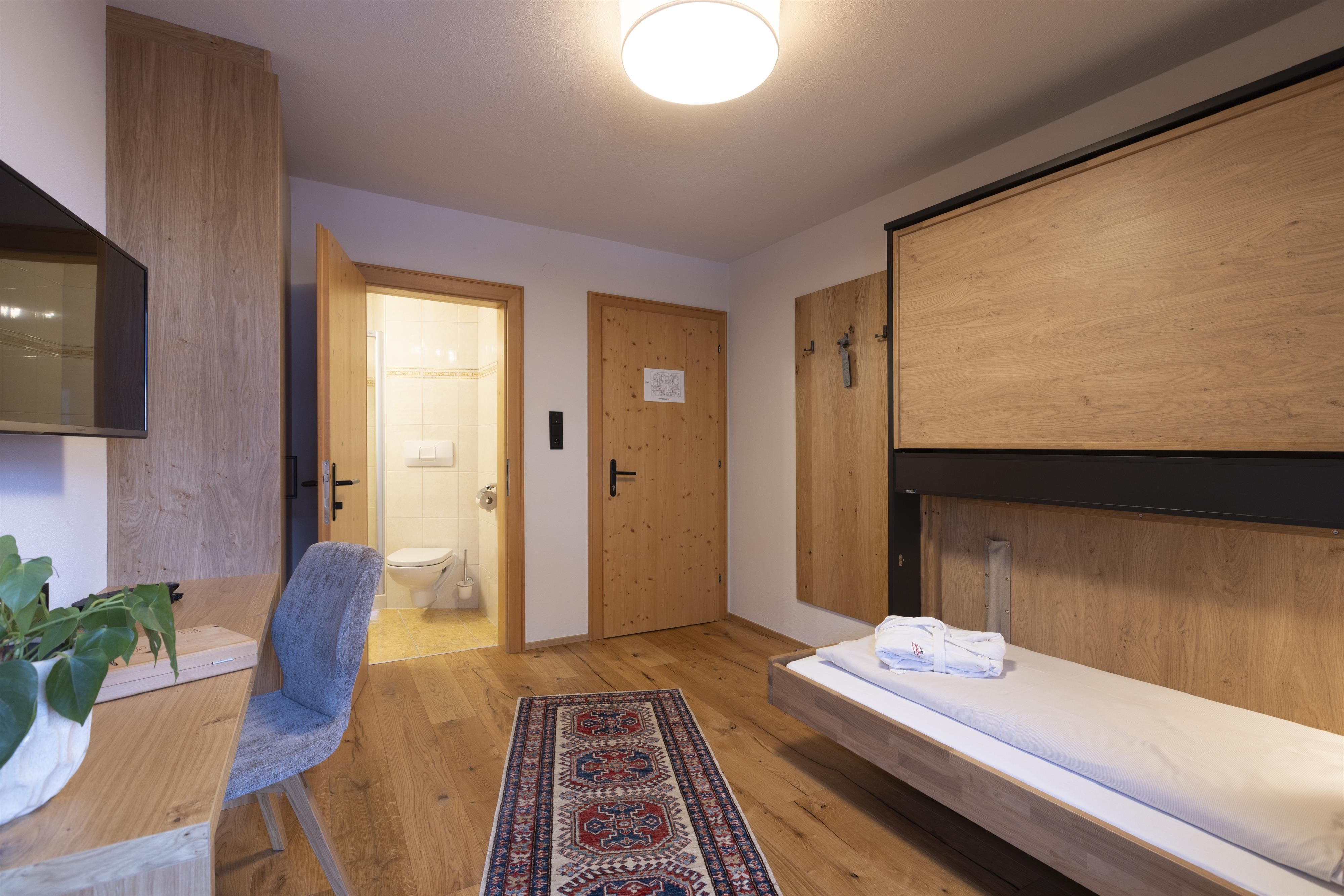 A modern room with wooden furniture and a color concept in natural tones. There is a desk, a television, and an adjacent bathroom.