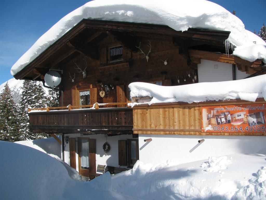A cozy wooden house with a snow-covered roof and balcony. The surroundings are enveloped in deep snow and the sun is shining.