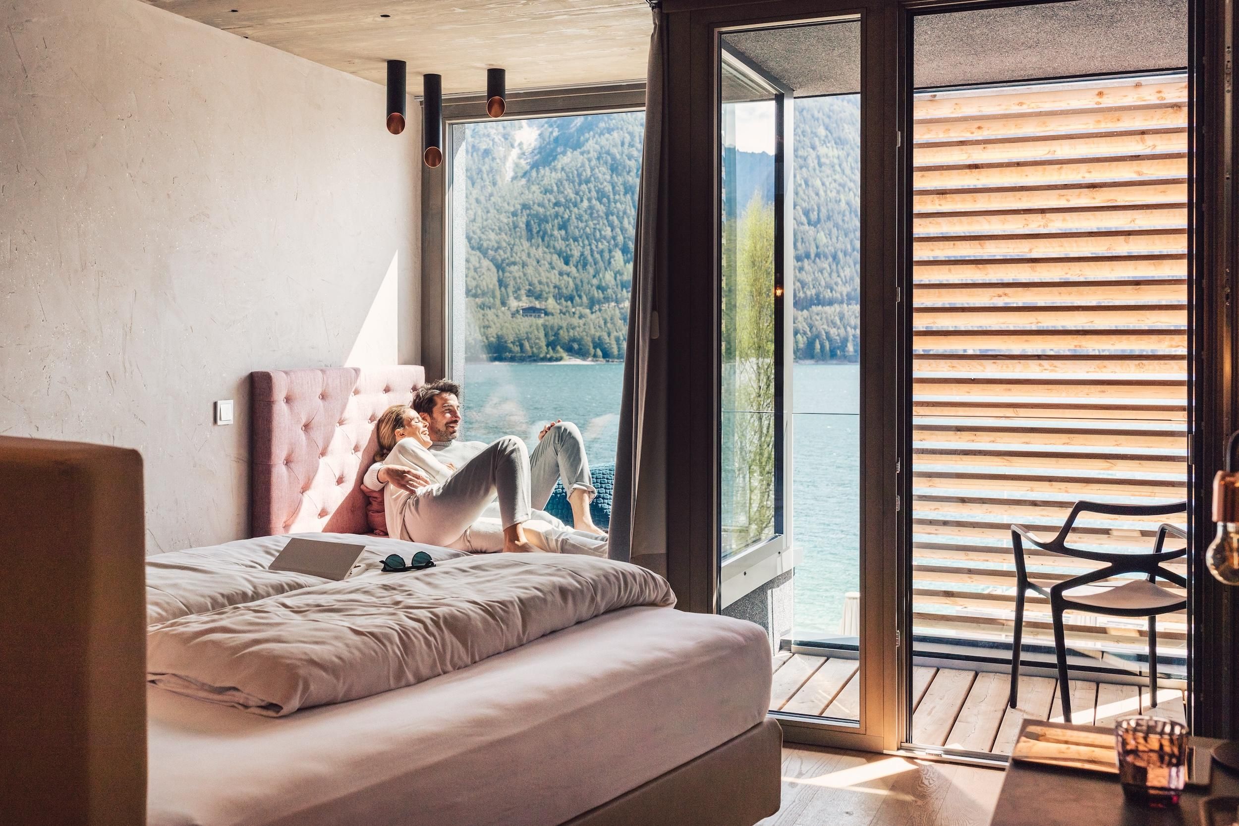 A modern room with a large bed and a balcony overlooking the lake. A couple is relaxing comfortably by the window and enjoying the view of the mountains.