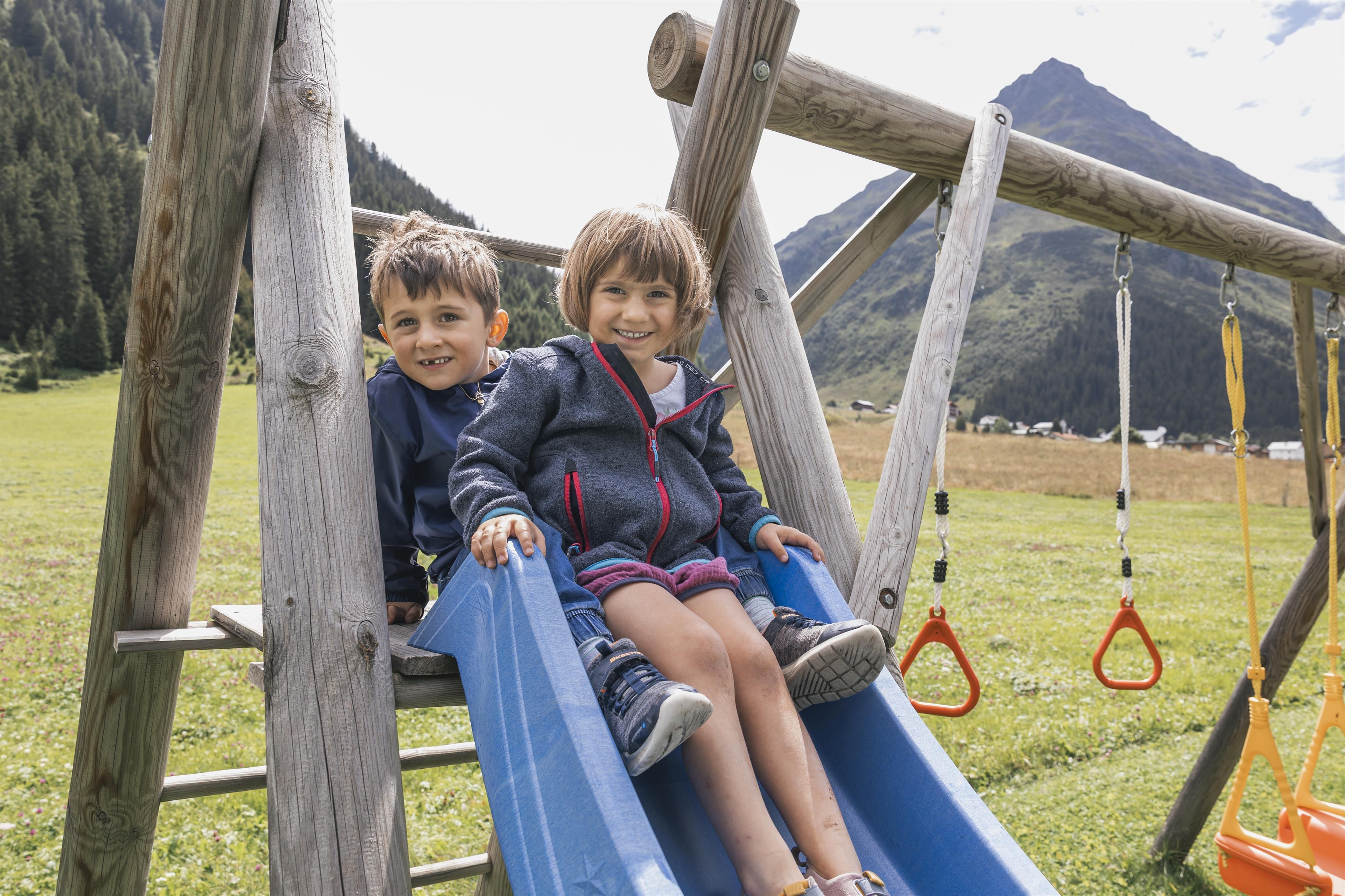 Two children are happily sitting on a slide in a playground. In the background, green meadows and mountains can be seen.