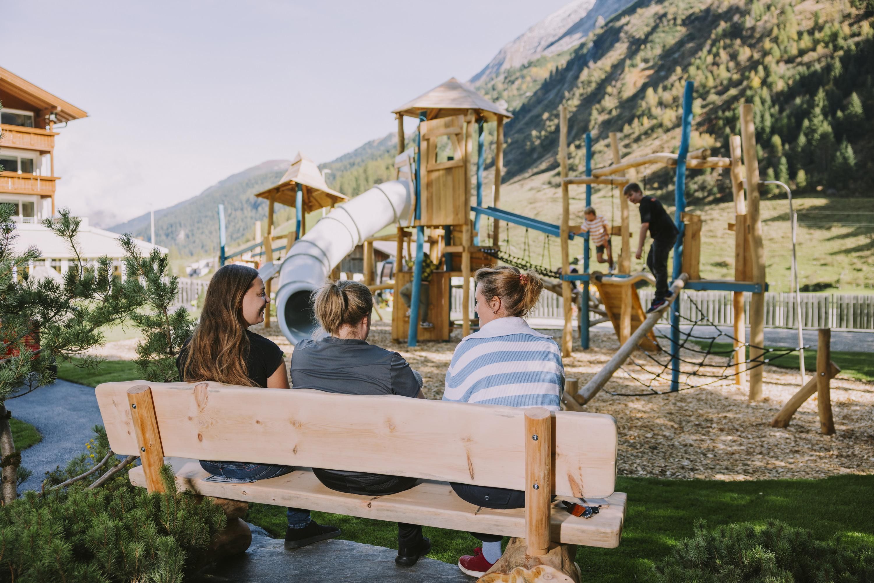 A playground in the mountains with wooden play equipment and a slide. In the foreground, three people are sitting on a bench watching the children.