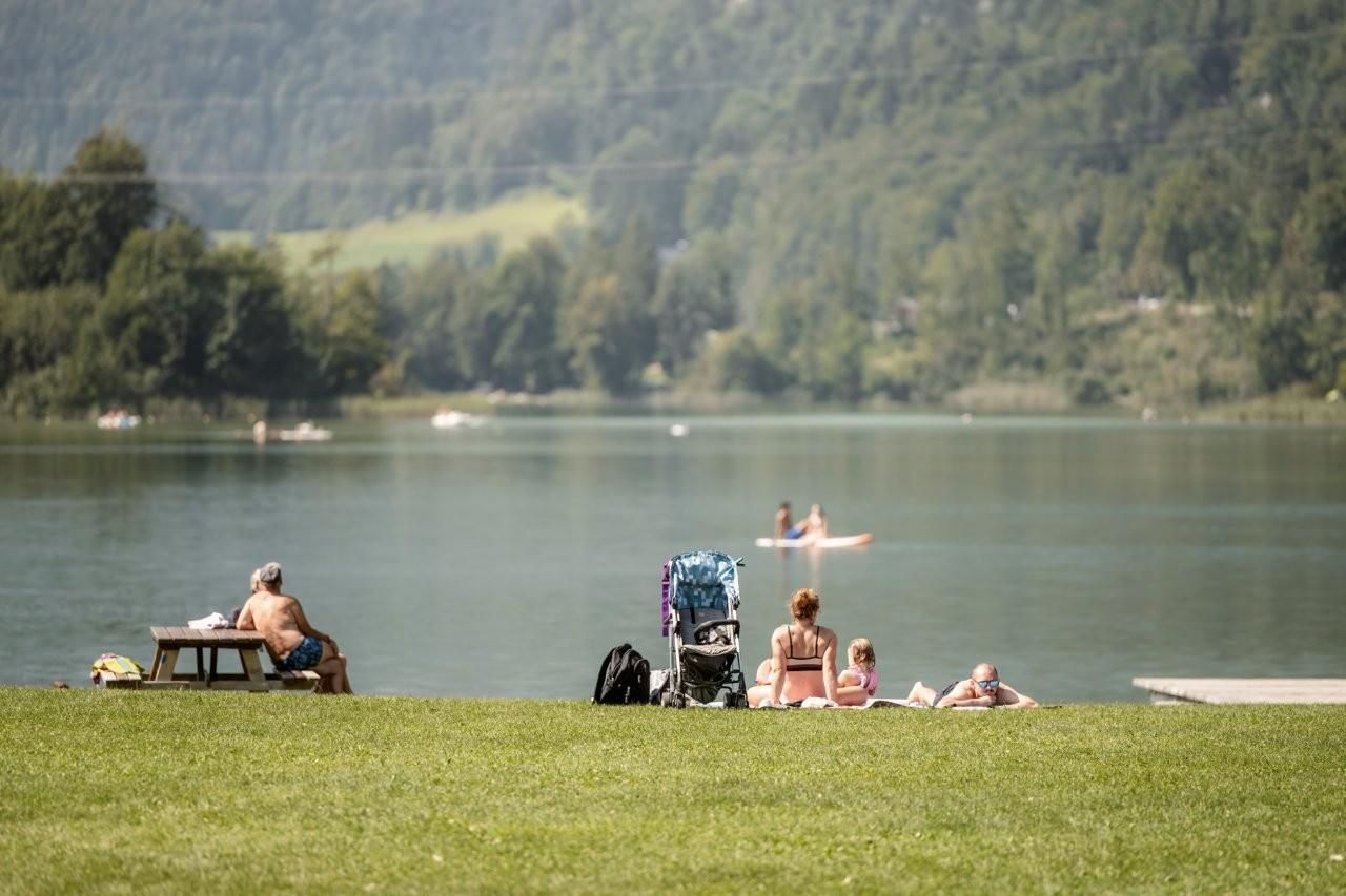 A calm lake with green meadows and people relaxing. In the background, a few individuals are paddling on the water.