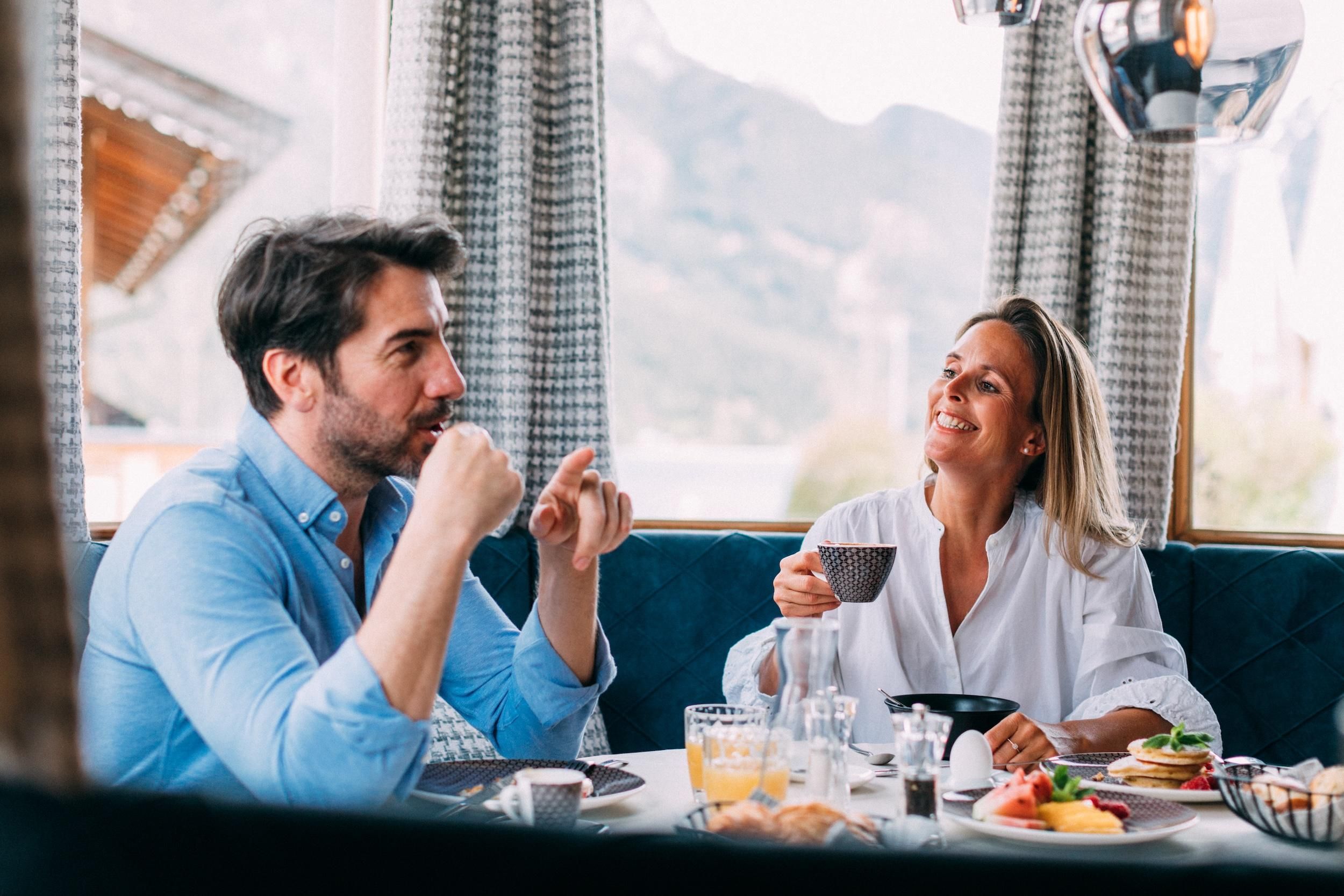 A couple is sitting at a table and enjoying breakfast. The atmosphere is relaxed and friendly with a view of the mountains.