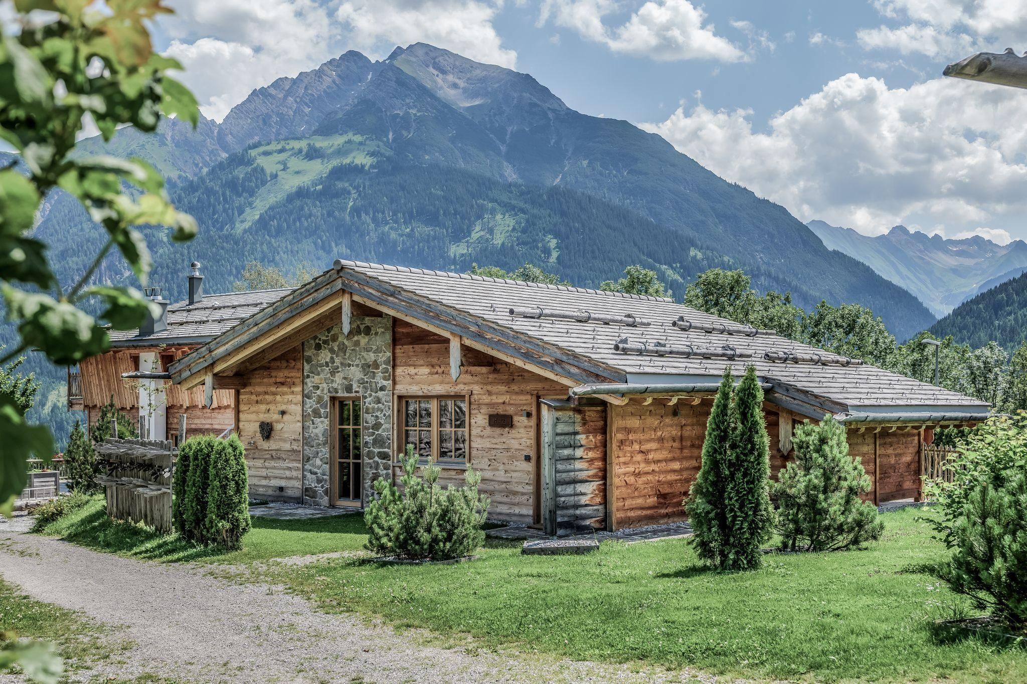 A cozy chalet surrounded by green meadows and mountains. The sky is partly cloudy and the landscape appears peaceful.
