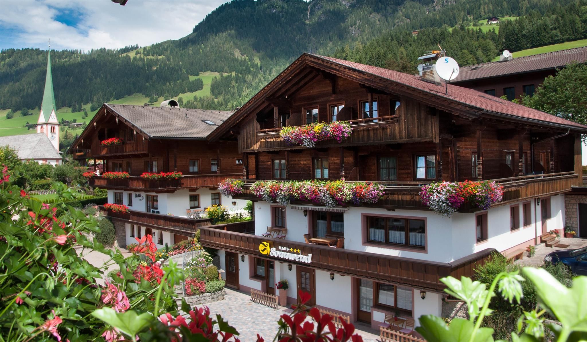 A charming Alpine house with a wooden facade and colorful flowers on the balconies. In the background, green mountains and a church tower can be seen.