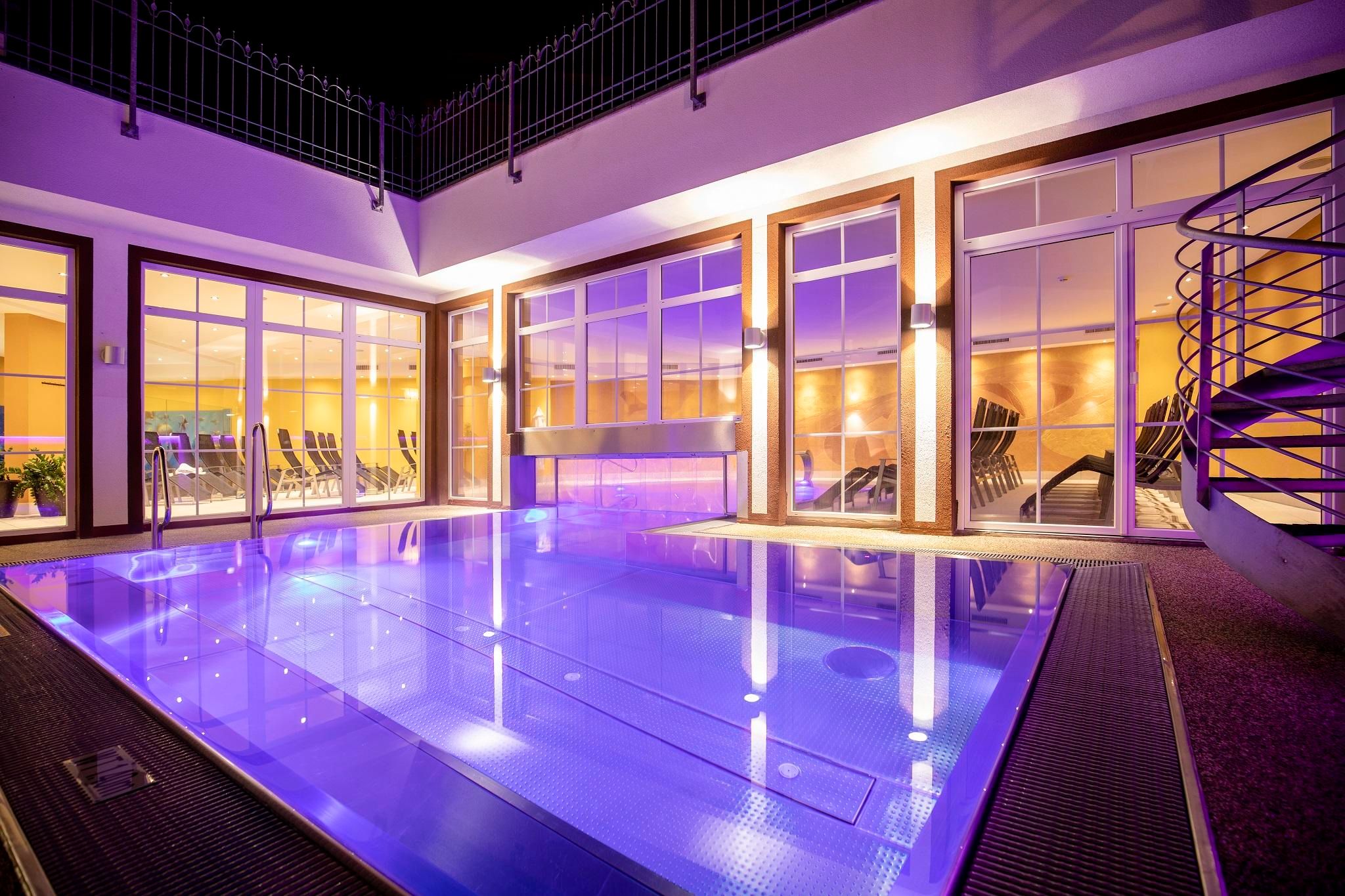 An elegant indoor pool with soft lighting. The large windows offer a view of a modern space with a moody atmosphere.