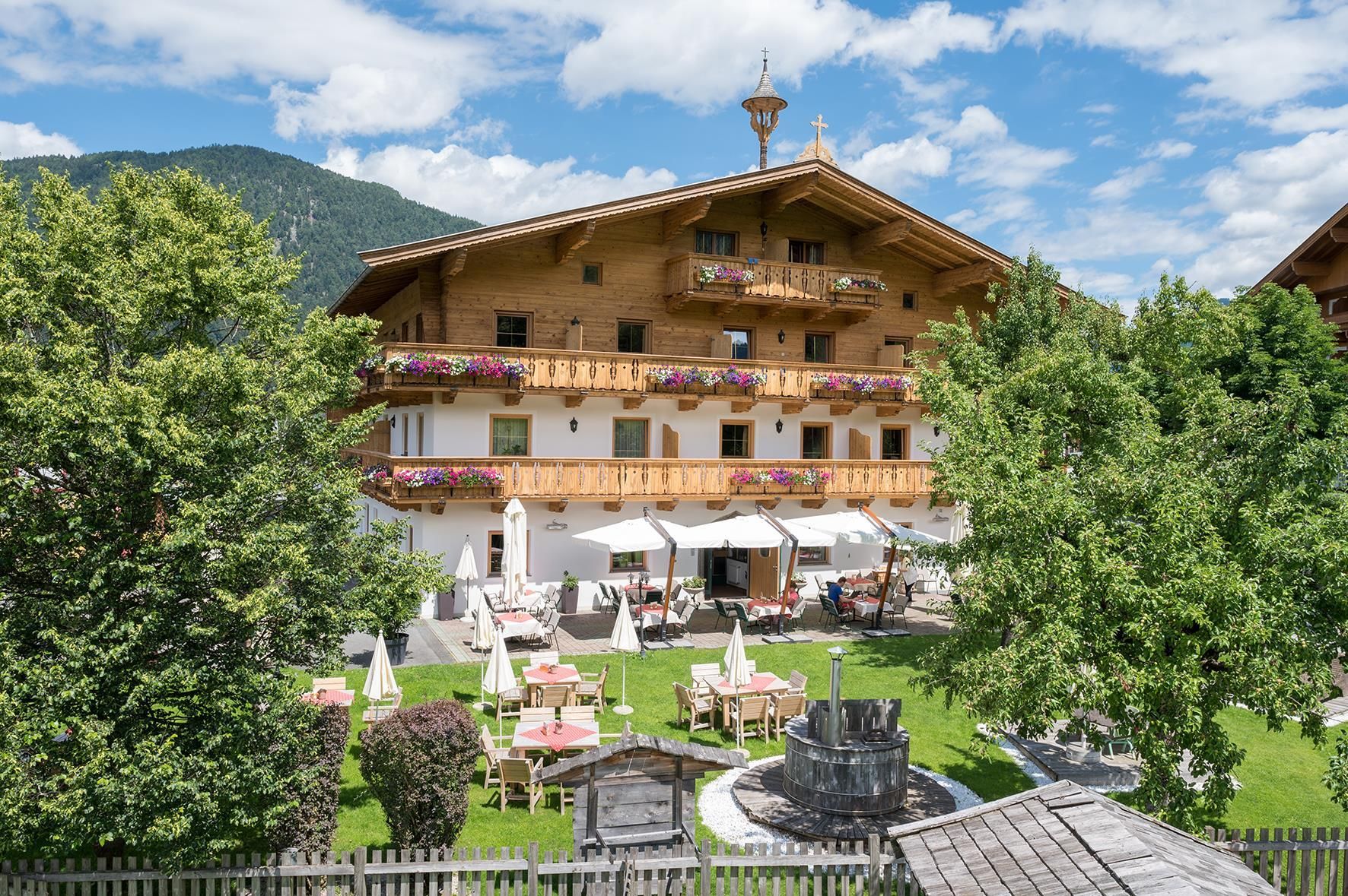 A traditional wooden building with balconies and flower boxes. In the foreground, there is a garden with lounge chairs and sun umbrellas.