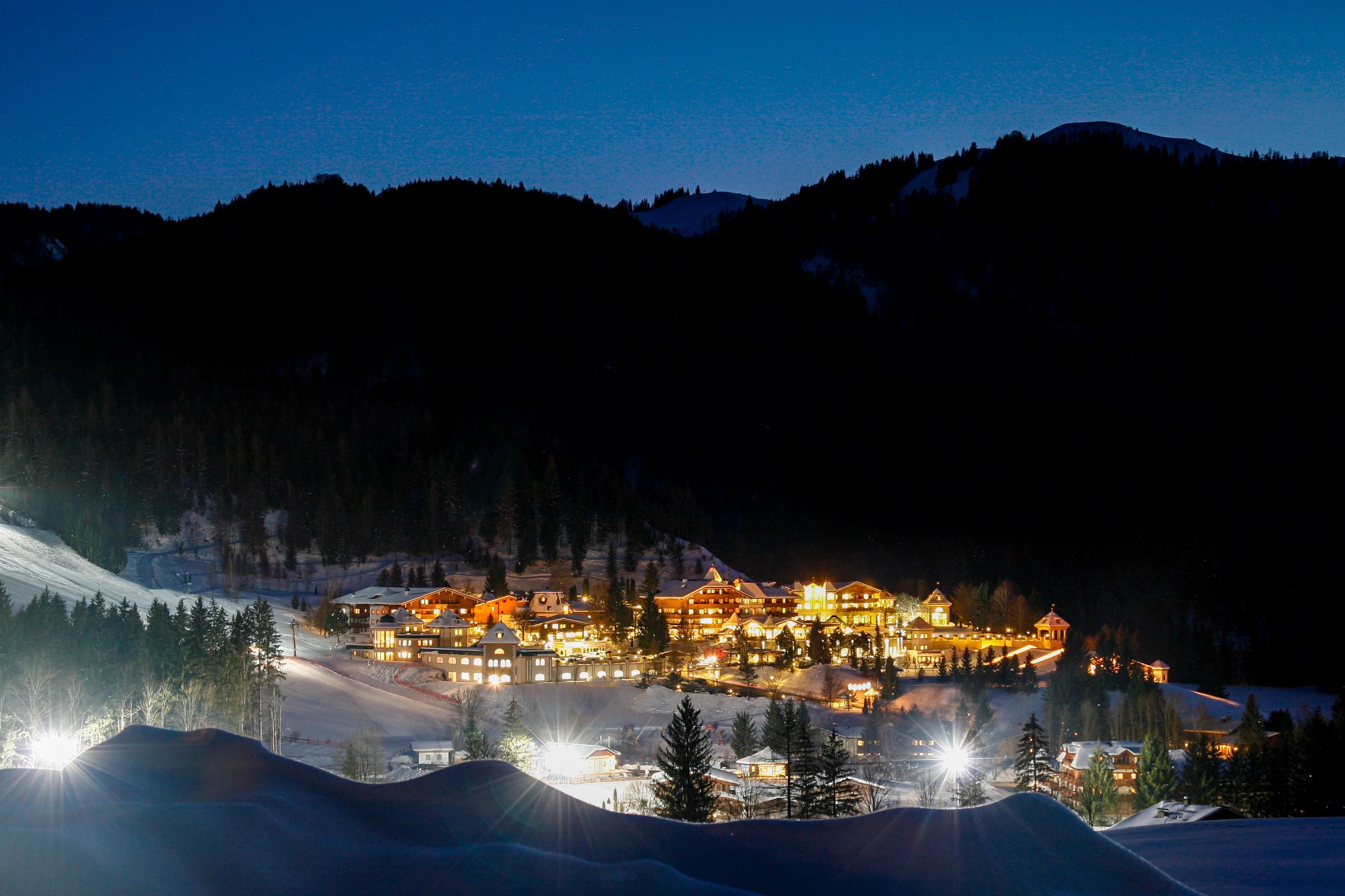 A picturesque mountain village at night, brightly lit by warm lights. In the foreground, there are snow-covered hills and forests.