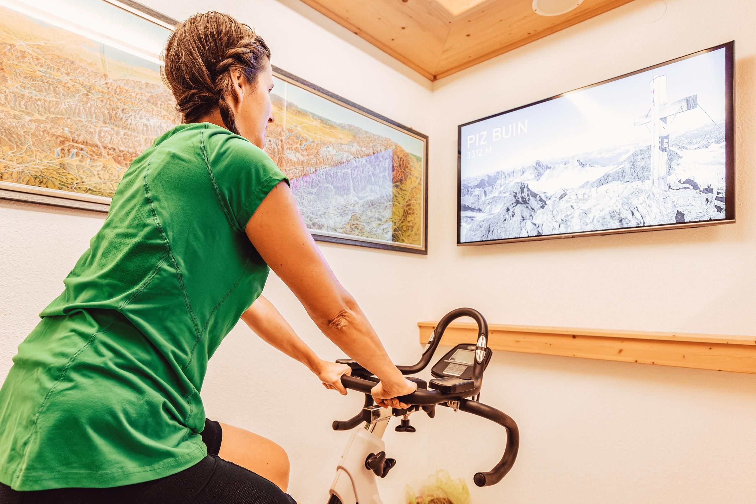 A person is riding on an exercise bike in a modern room. In the background, there is a screen with landscape footage.