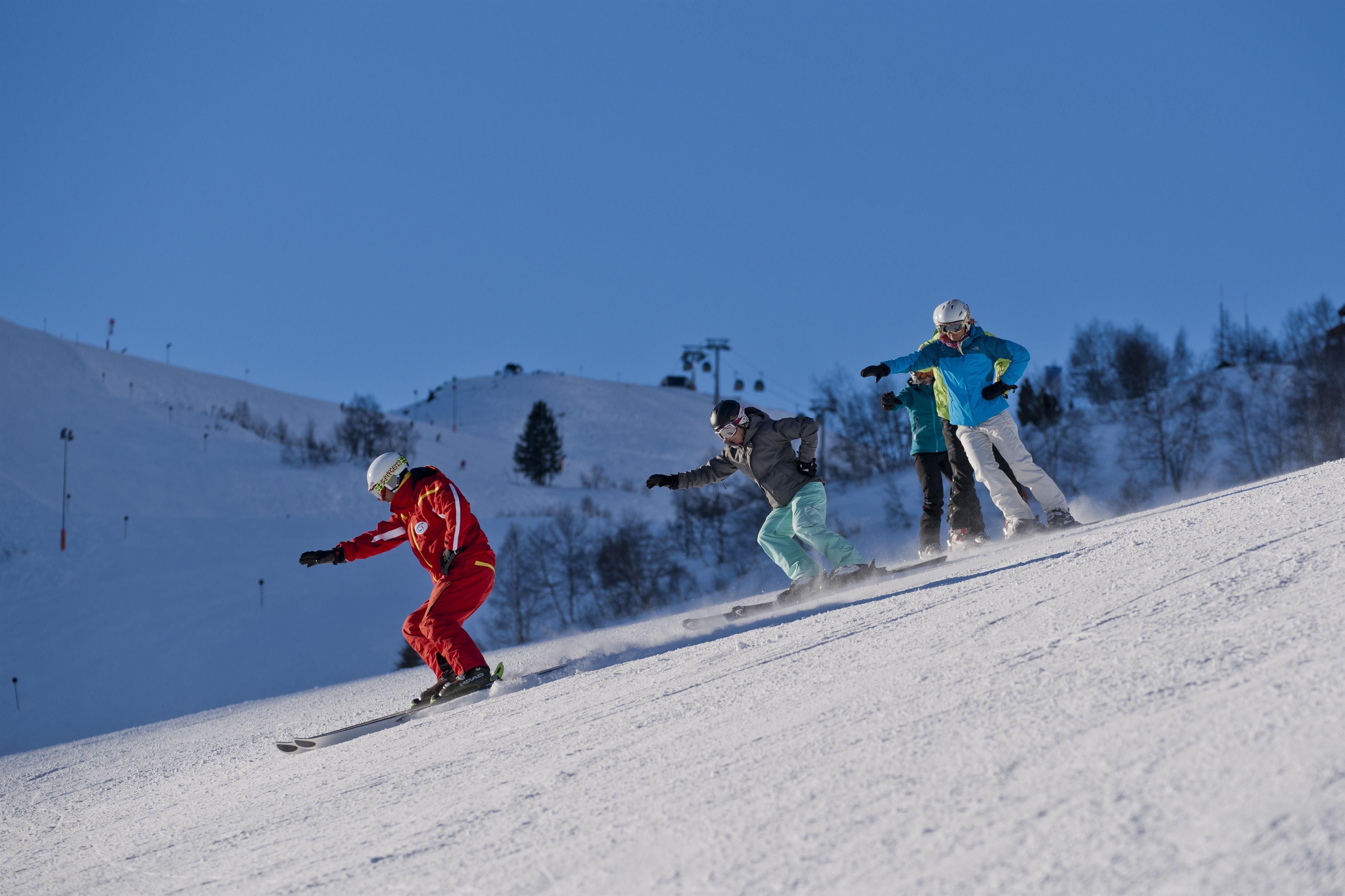 Three skiers go down the slope. The surroundings are snow-covered and the sky is clear and blue.
