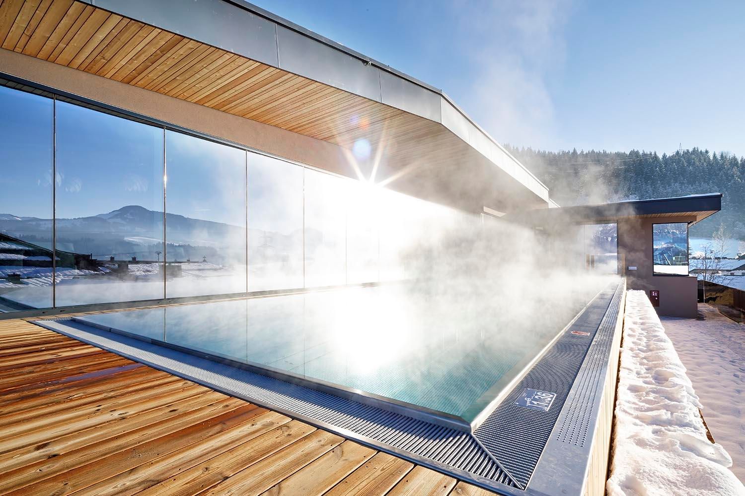 A modern pool with steam clouds, surrounded by glass and wood. The view of the mountains is clear and impressive.