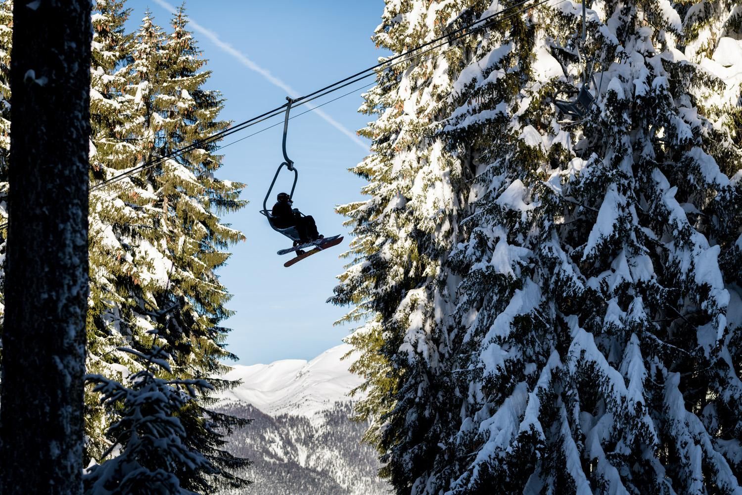 A skier rides a chairlift through a snow-covered forest landscape. In the background, mountains and a clear blue sky can be seen.
