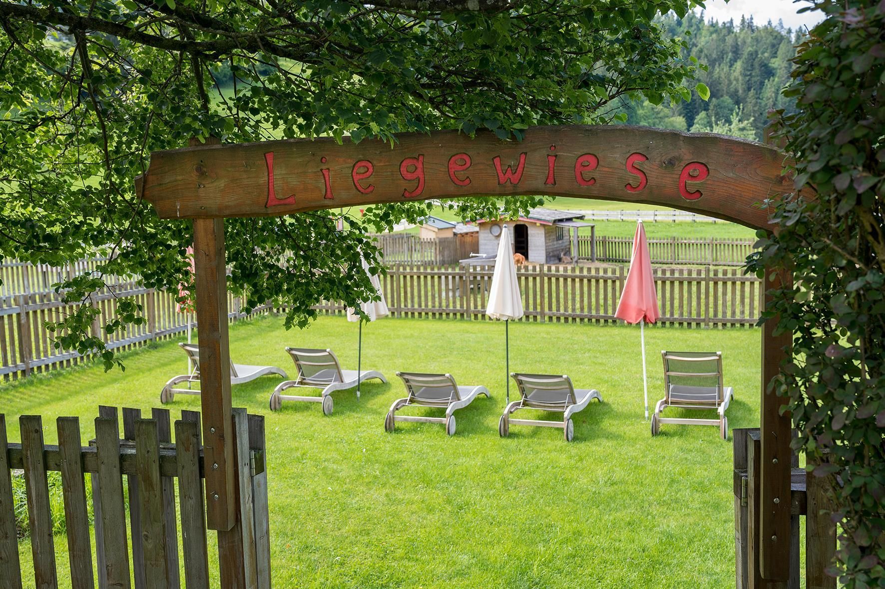A sunbathing area with loungers and sun umbrellas in a green garden. In the background, trees and a fenced area can be seen.