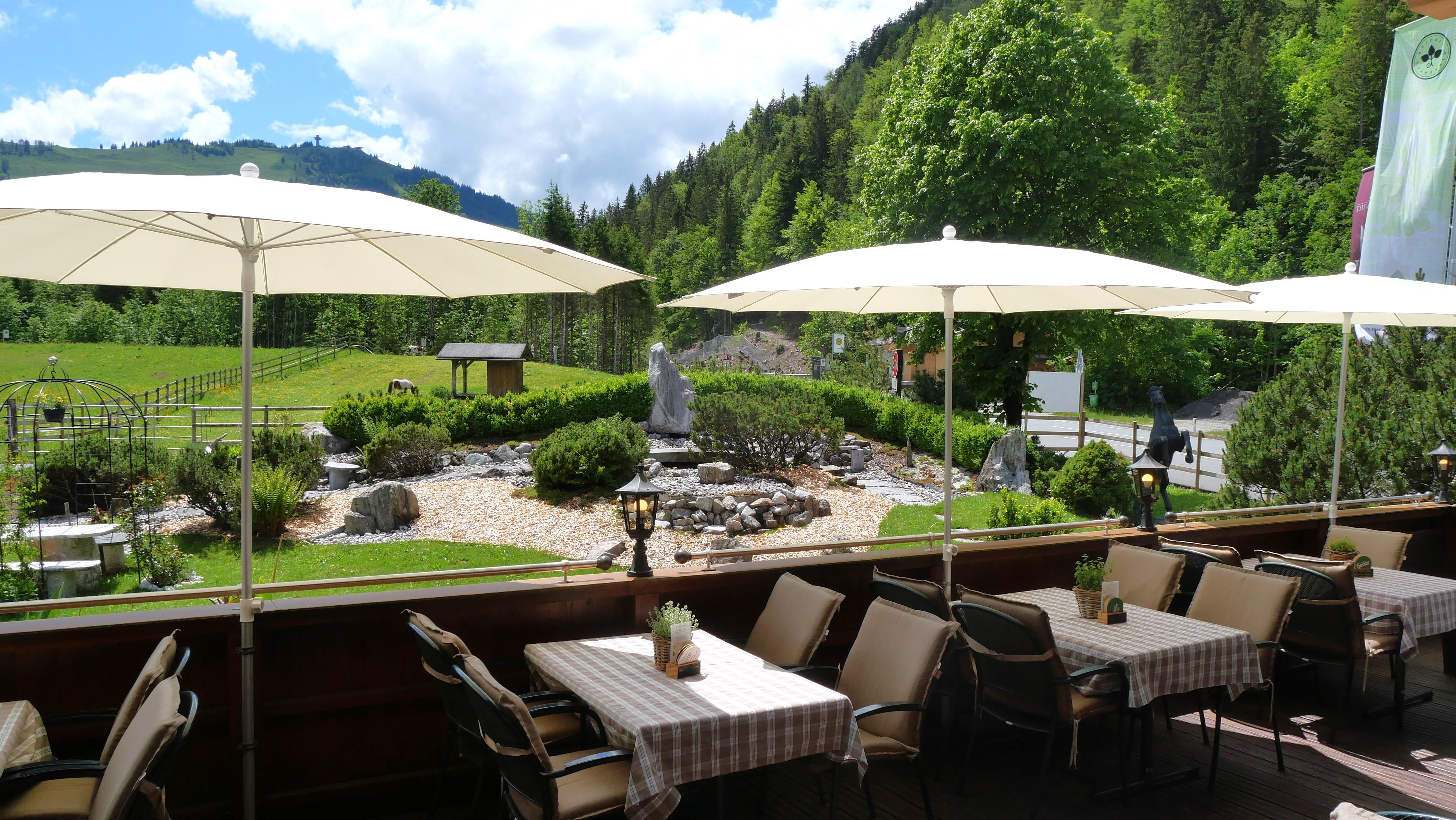 A cozy terrace with tablecloths and sun umbrellas. In the background, green trees and a well-maintained garden landscape can be seen.