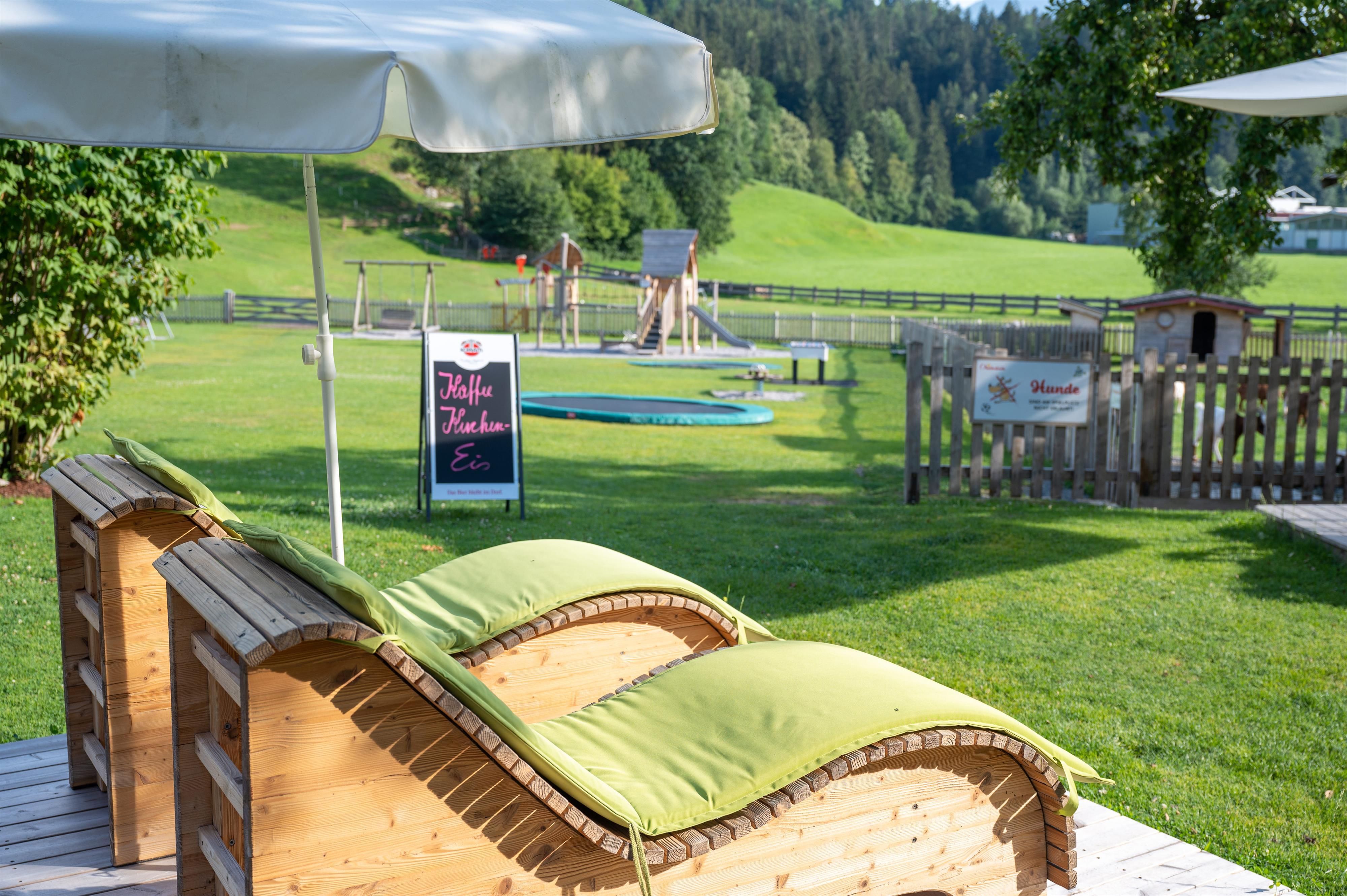 A tranquil garden landscape with loungers and sun umbrellas. In the background, there are play areas for children and a green lawn.