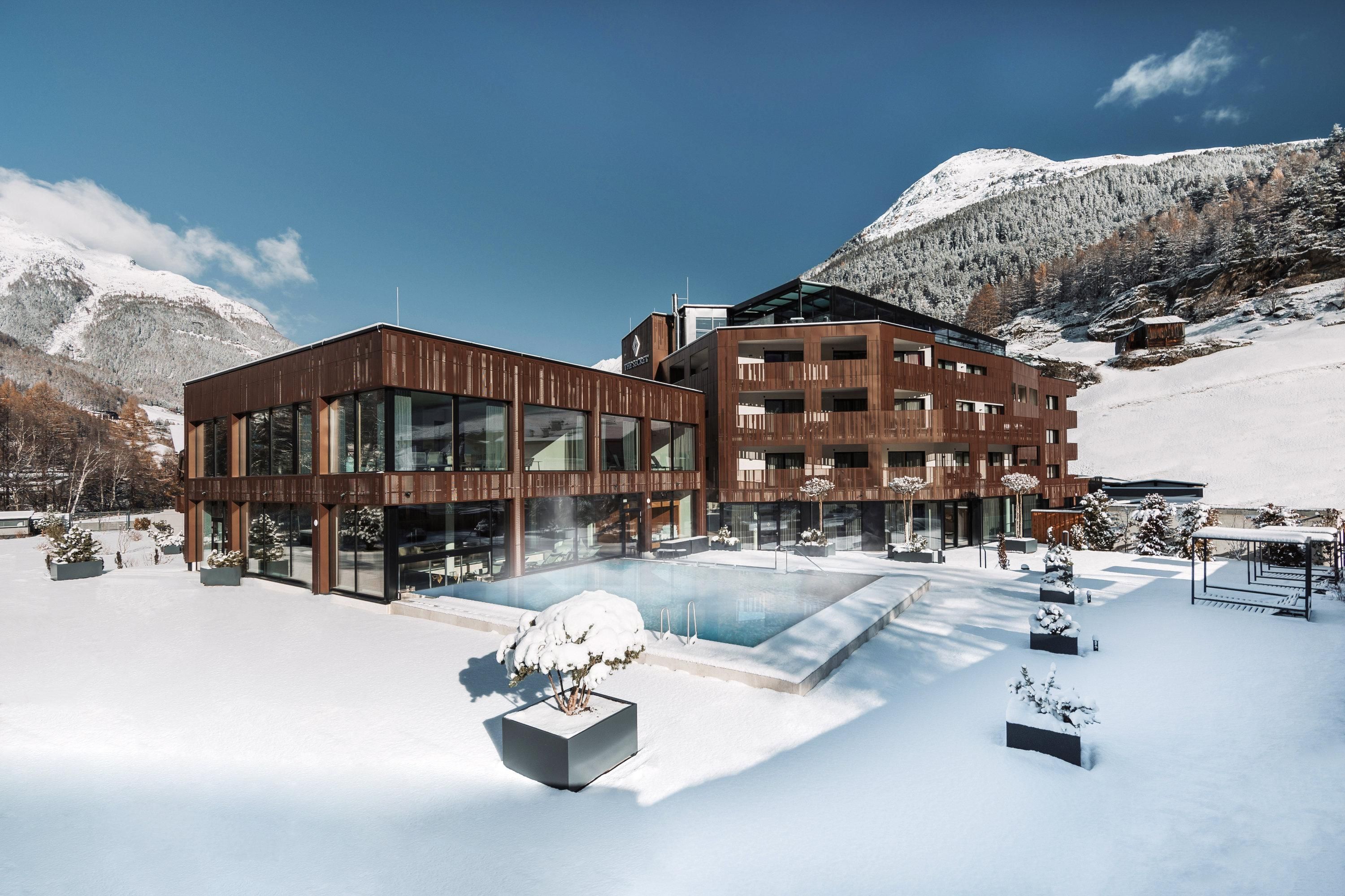 A modern hotel in the snow with an outdoor spa and surrounding mountains. The facility is surrounded by a winter landscape.