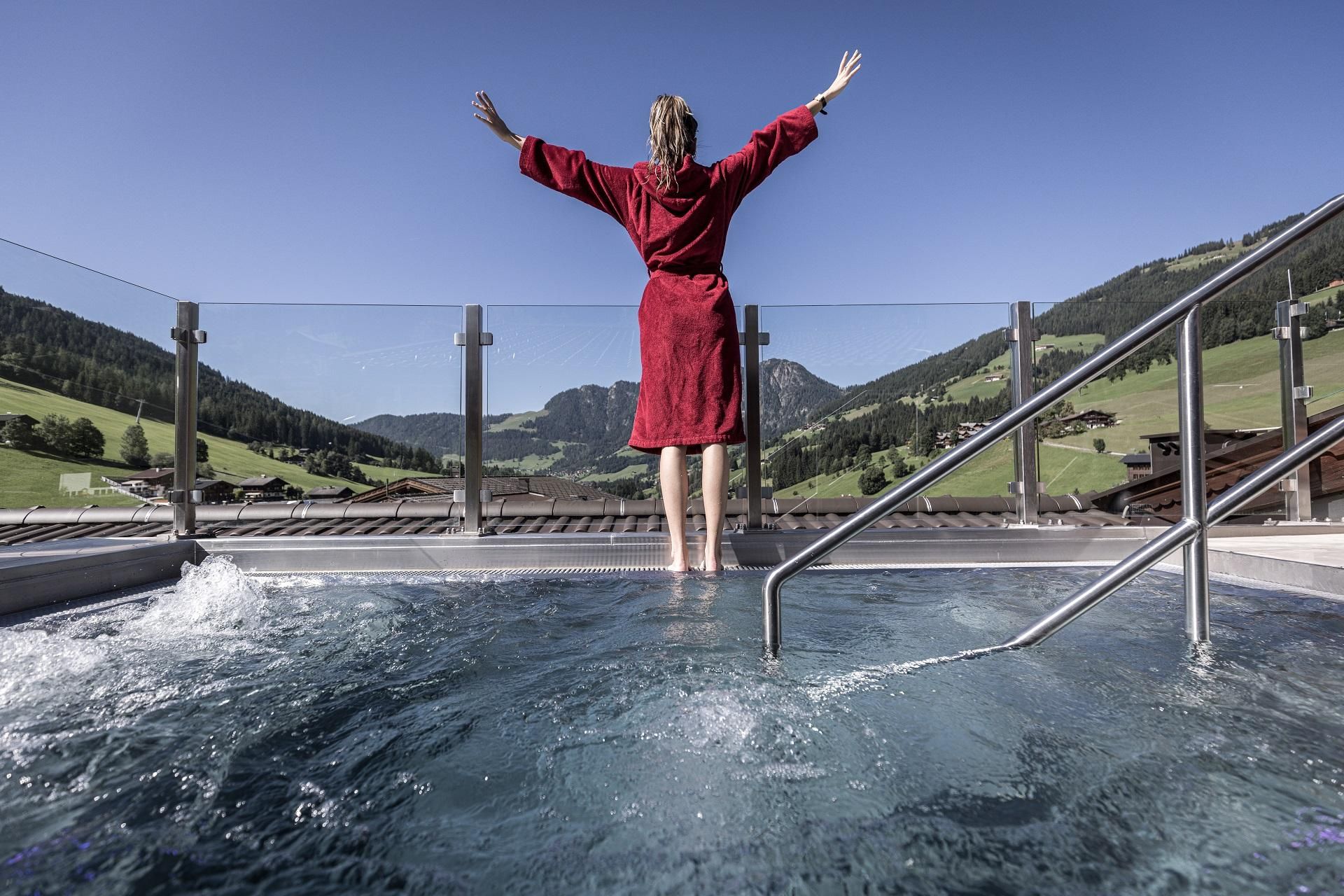 A person in a red bathrobe stands at the edge of a hot tub. In the background, a picturesque mountain landscape can be seen under a clear sky.
