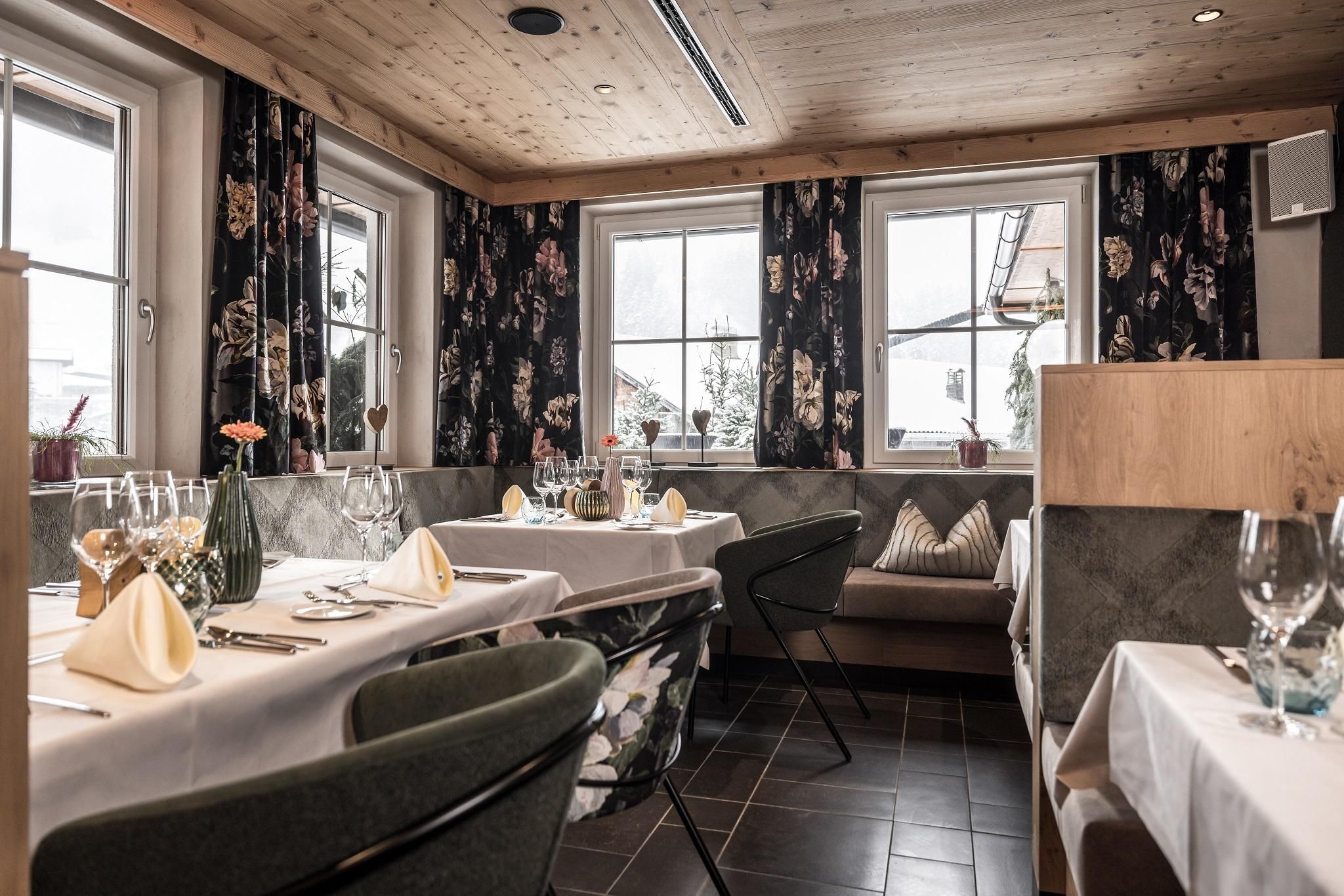 An elegant restaurant with a wooden ceiling and stylish floral curtains. The tables are tastefully set, creating an inviting atmosphere.