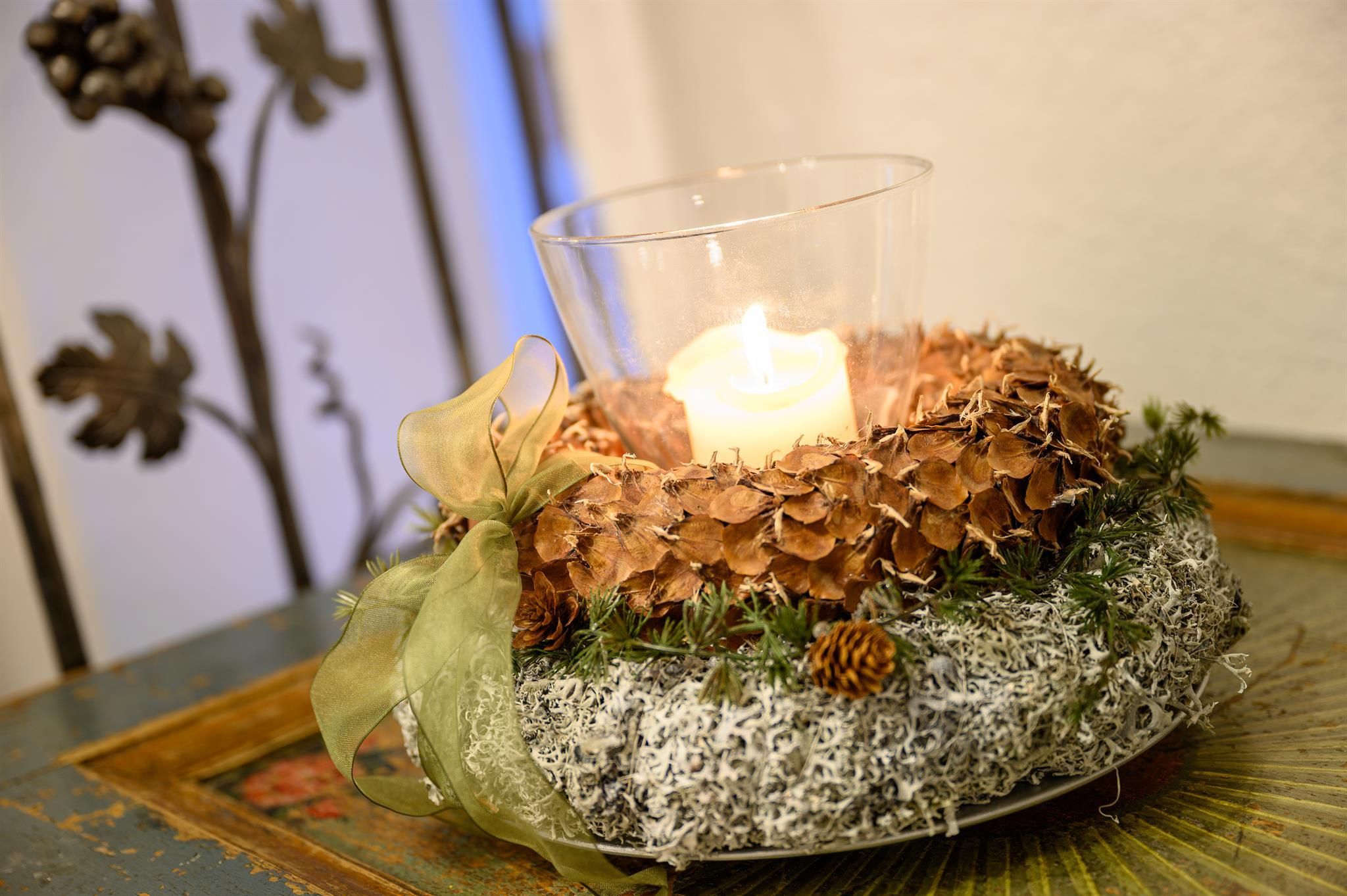 A decorative wreath made of pine cones and moss with a candle in the center. The wreath is adorned with a bow and creates a cozy atmosphere.