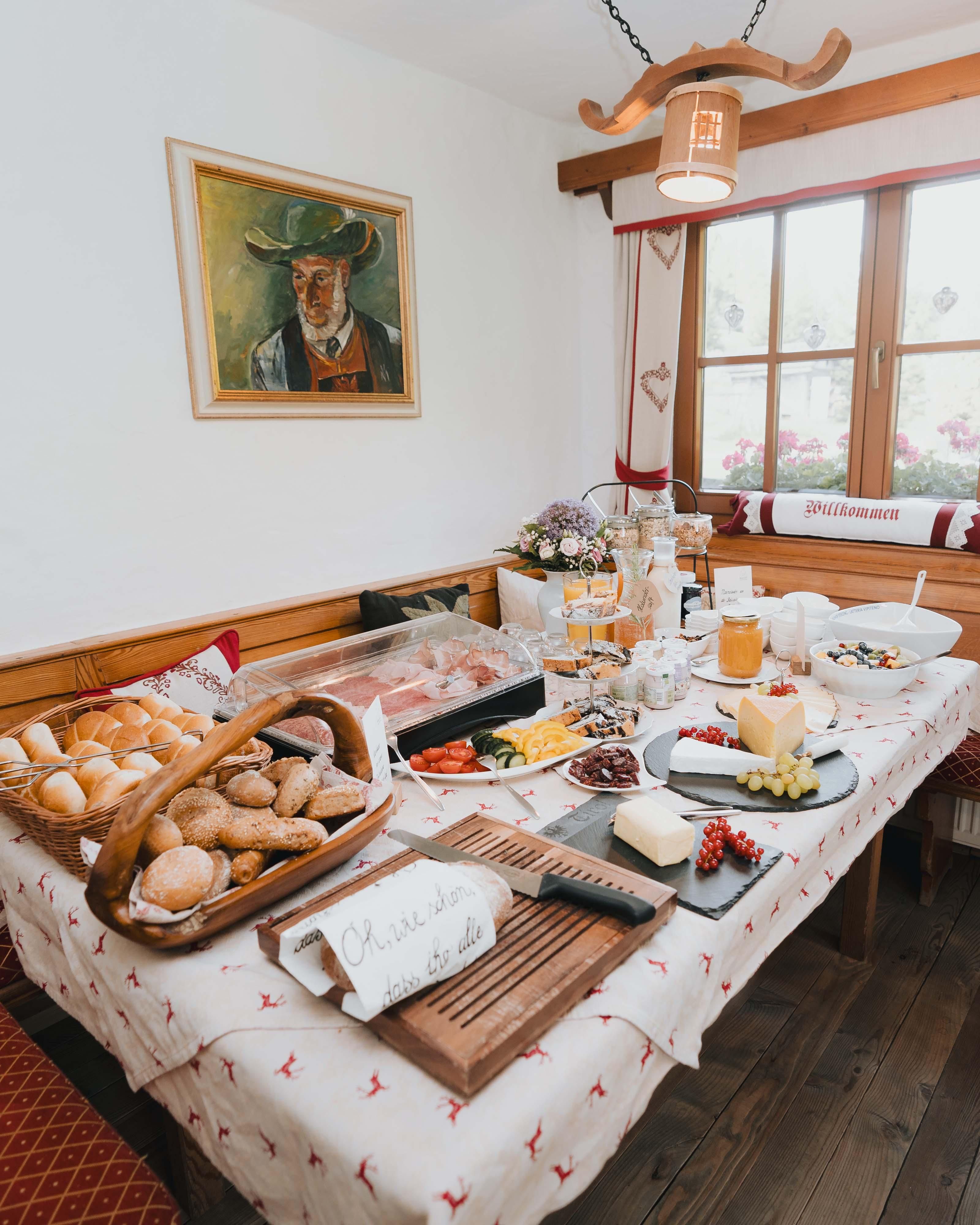 A set table with a selection of bread, cheese, fruit, and drinks. In the background is a painting and a window with a view outside.