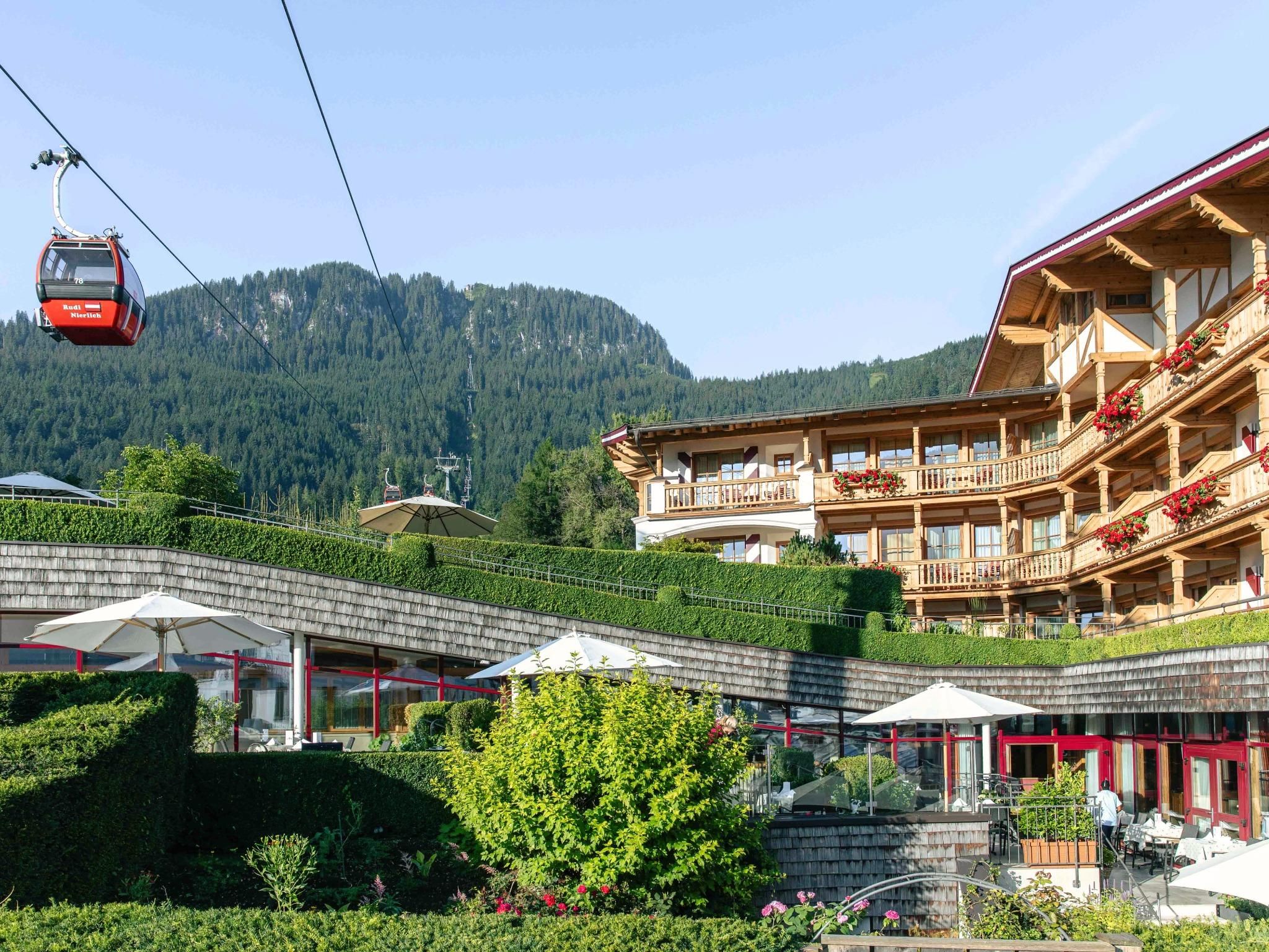 A picturesque mountain landscape with a red chairlift and a rustic building. The surroundings are green with well-maintained gardens and sunshades.