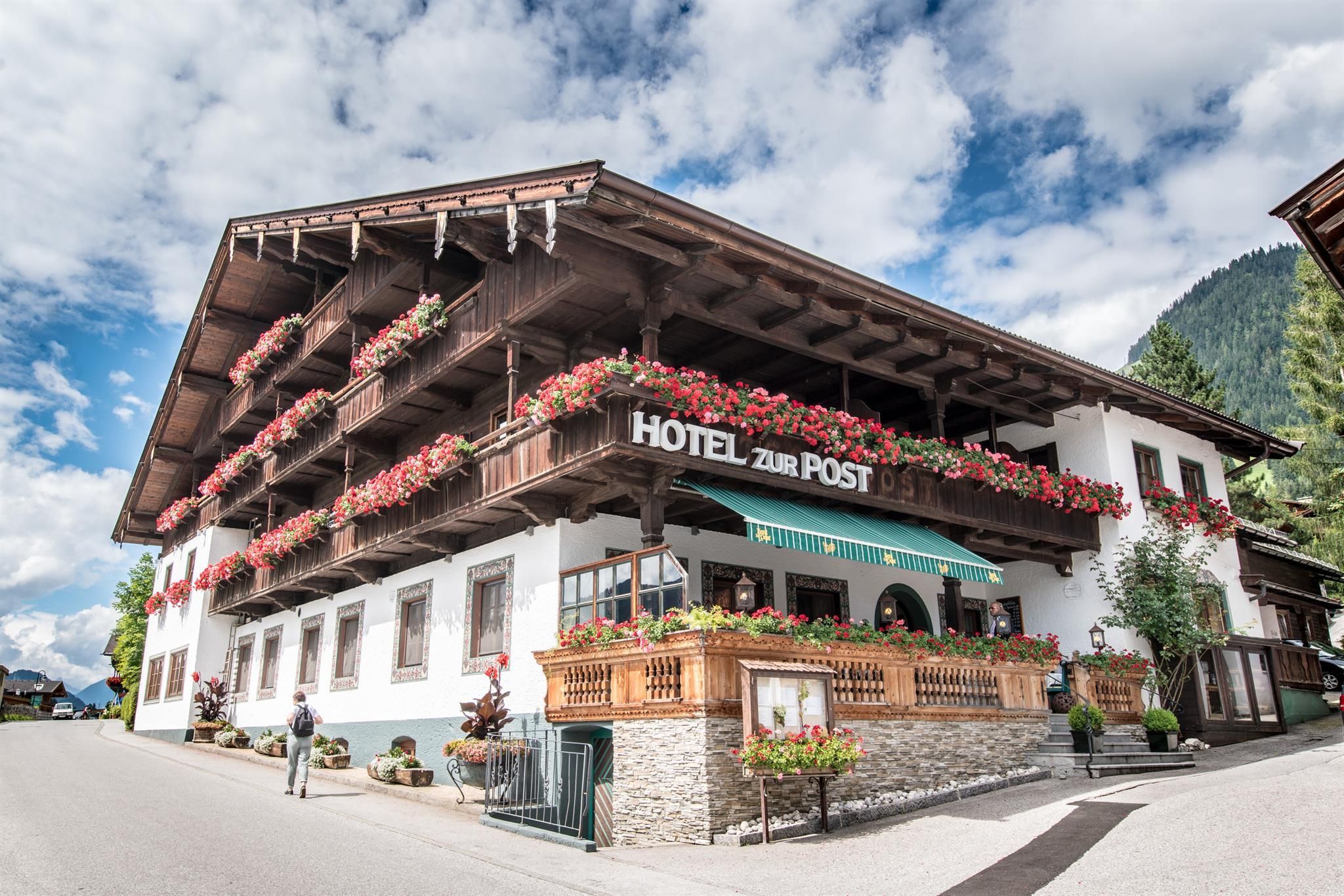 A traditional hotel with a beautiful wooden terrace and colorful flowers. The sky is clear and the surroundings are surrounded by mountains.