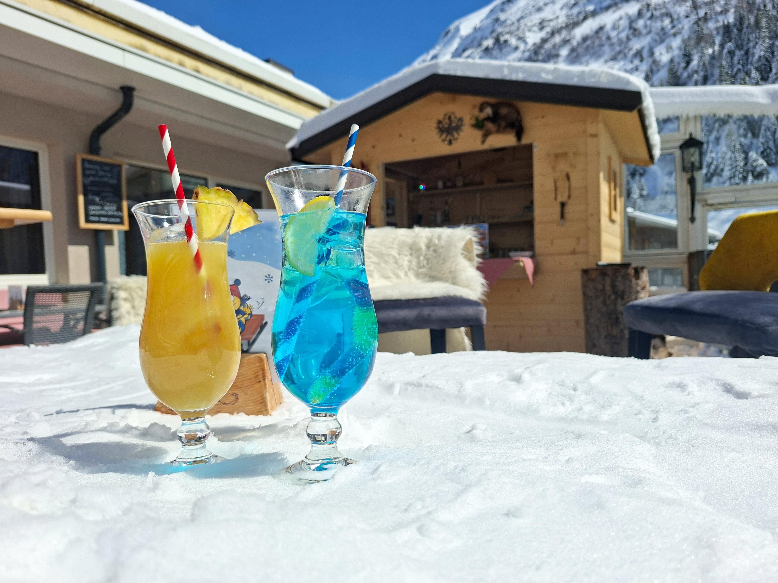 Two colorful cocktails are sitting on the snow. In the background, a chalet cabin and tall mountains can be seen.