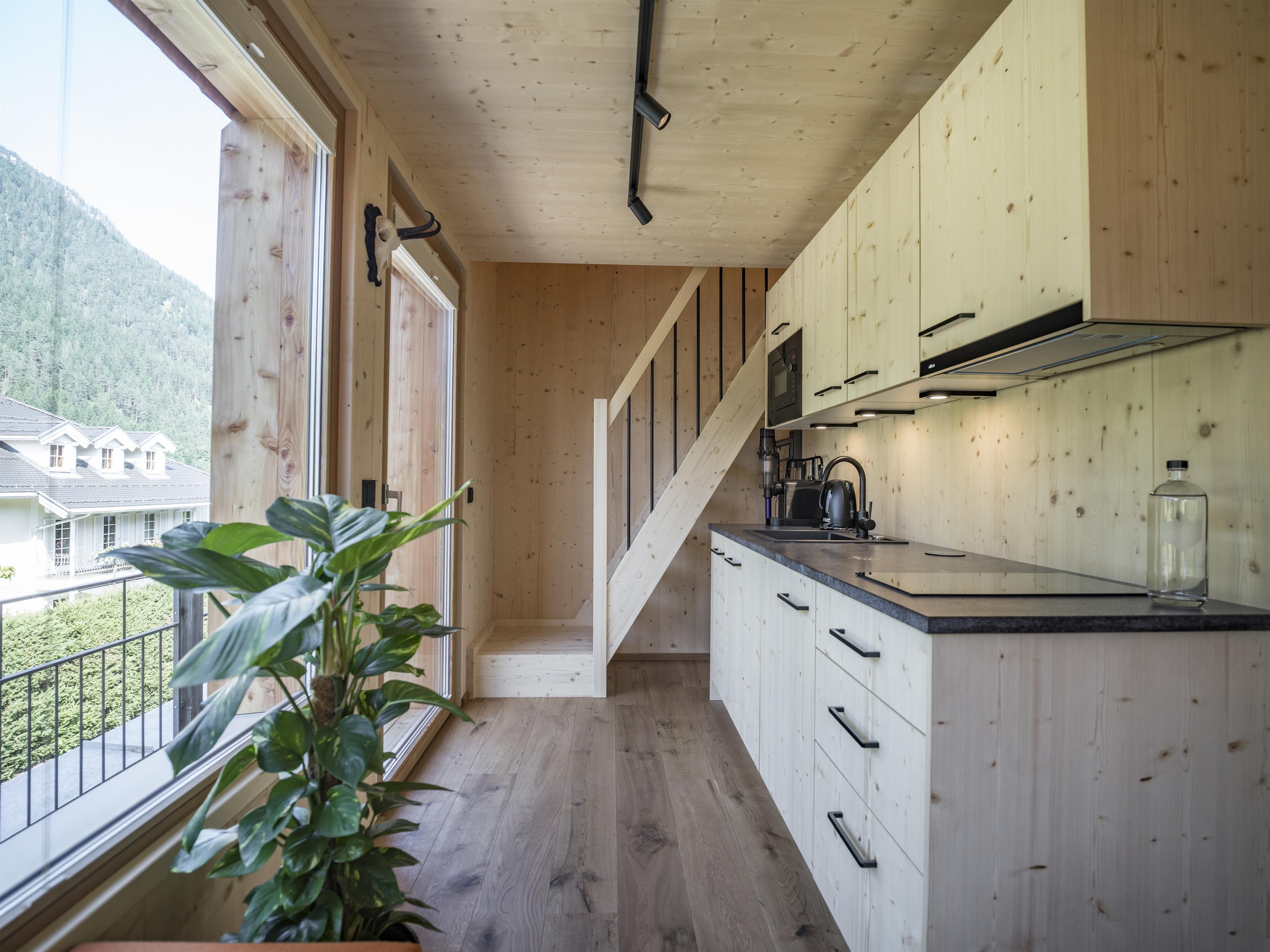A modern kitchen unit with wooden cladding and plenty of daylight. In the background, a staircase and a green plant can be seen.