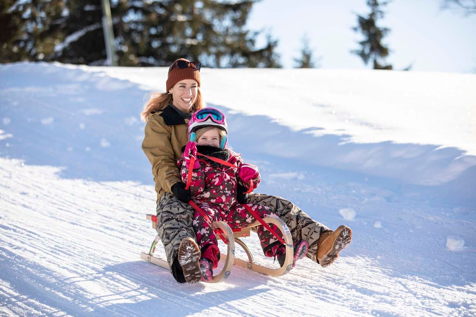 A mother and her child are riding together on a sled in the snow. The shining sun and the snowy landscape create a cheerful winter atmosphere.