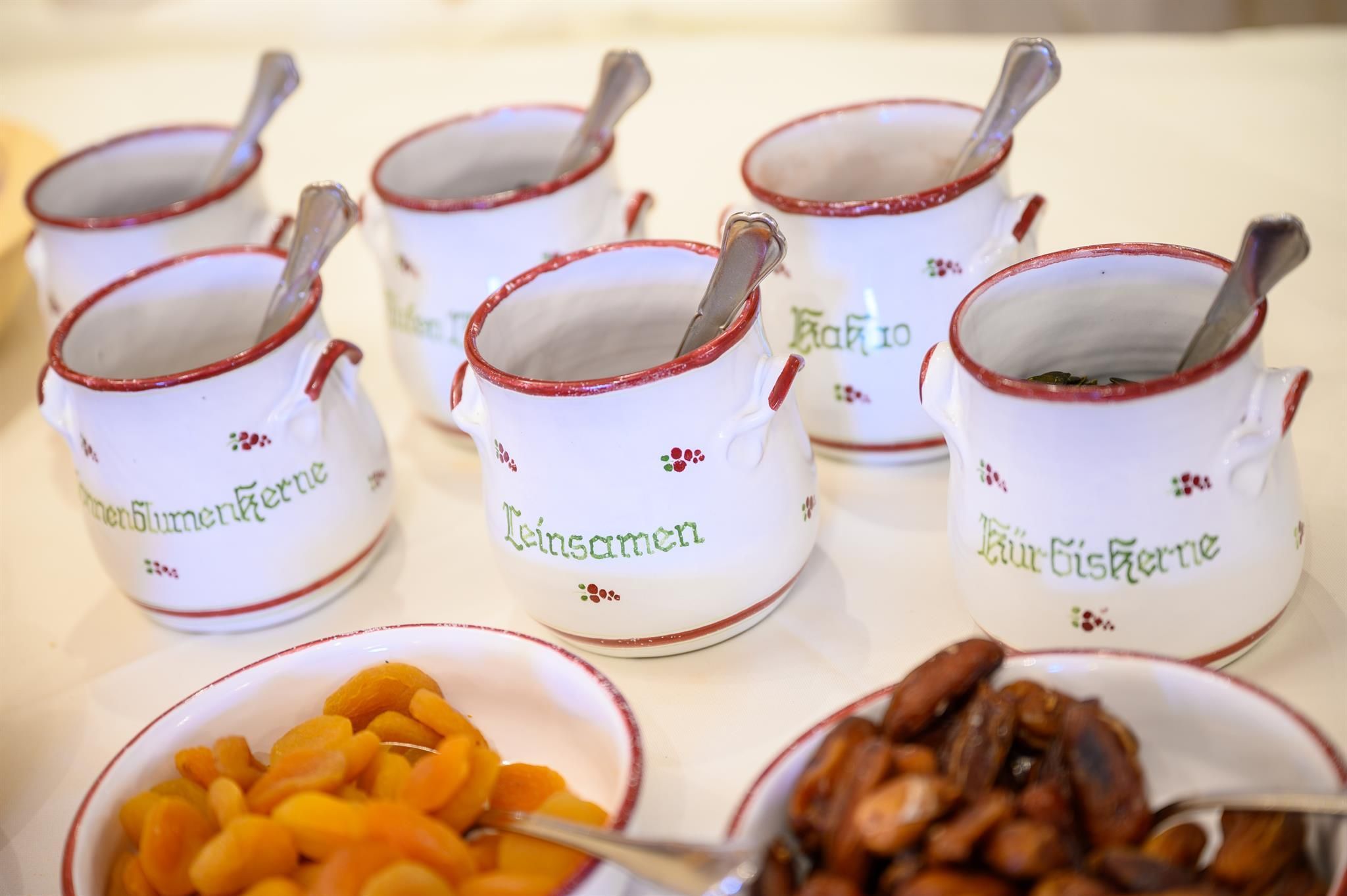 Six small pots with different ingredients are on a table. Next to them are dried apricots and nuts.
