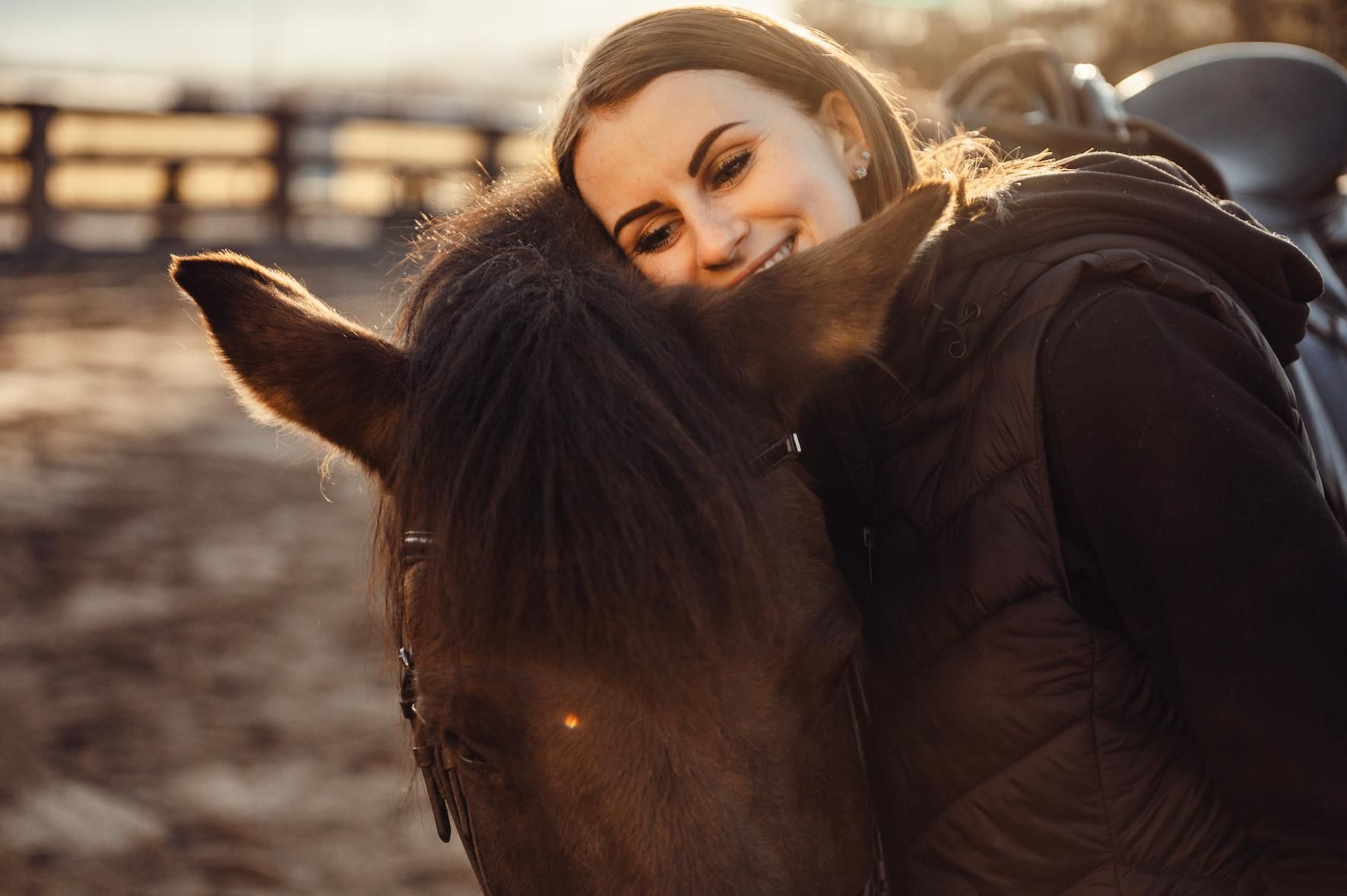 A woman hugs a black horse and smiles happily. The background shows a rural stable and warm sunlight.