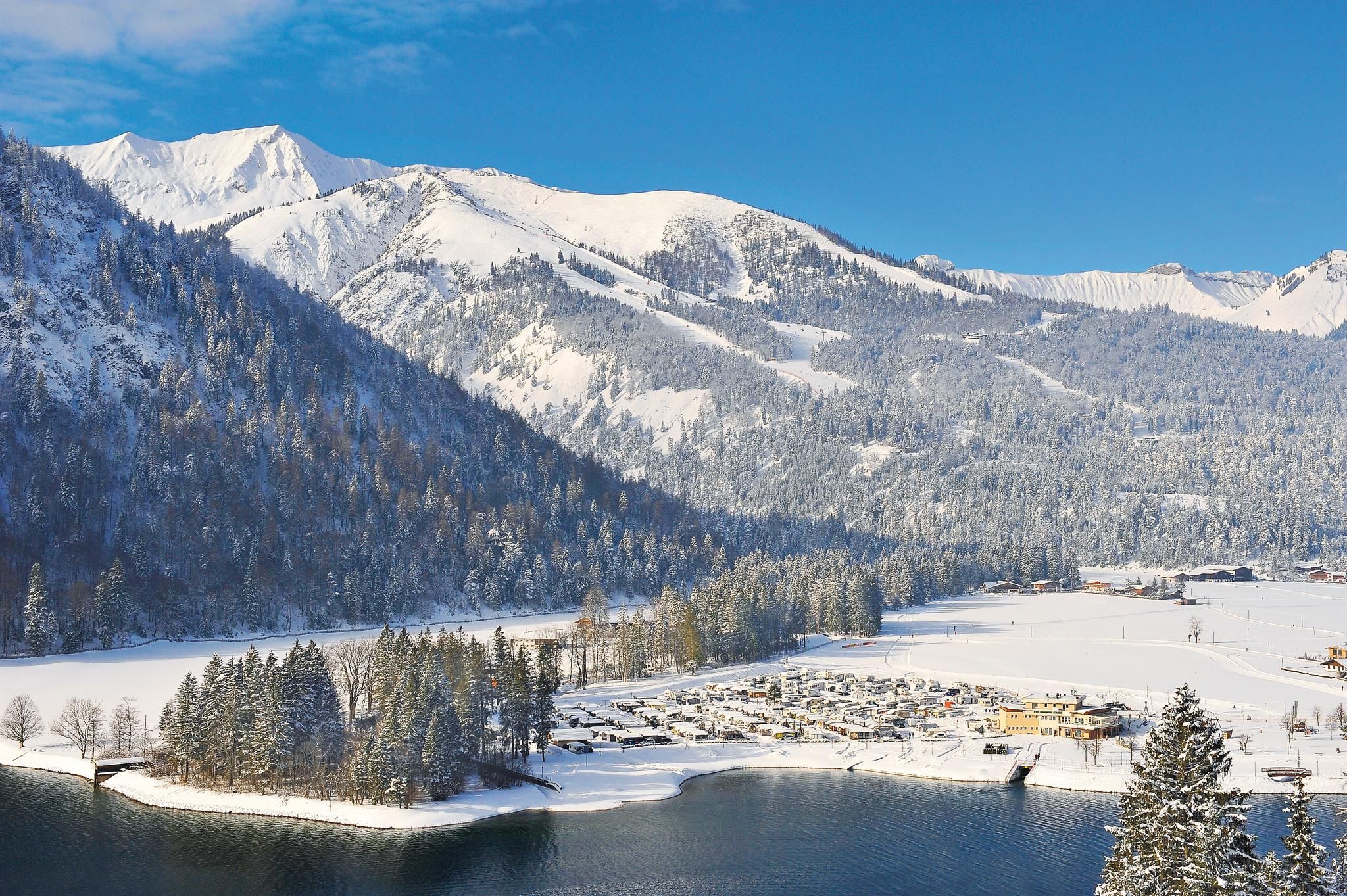 A snow-covered landscape with mountains and a serene lake. The sky is clear and blue, highlighting the winter beauty.