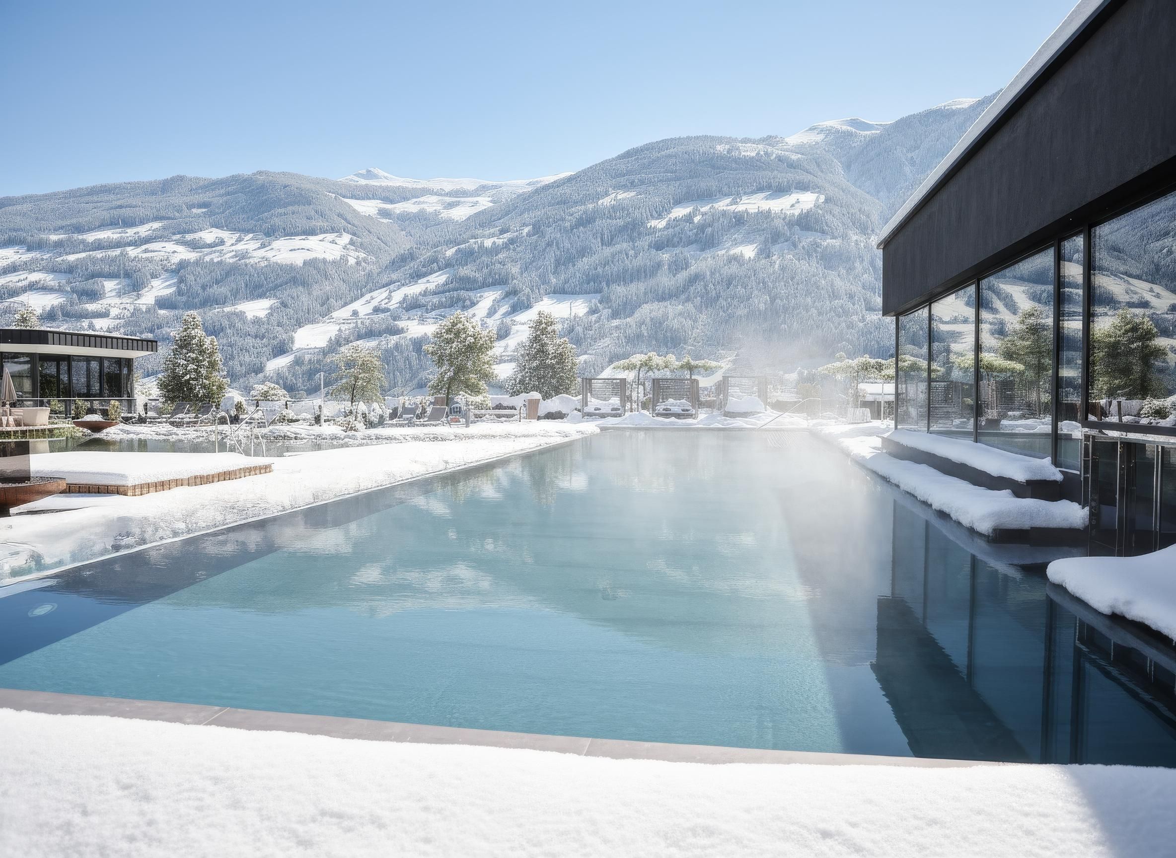 An elegant outdoor pool in a snow-covered setting with mountains in the background. The clear, calm water surface reflects the blue sky.