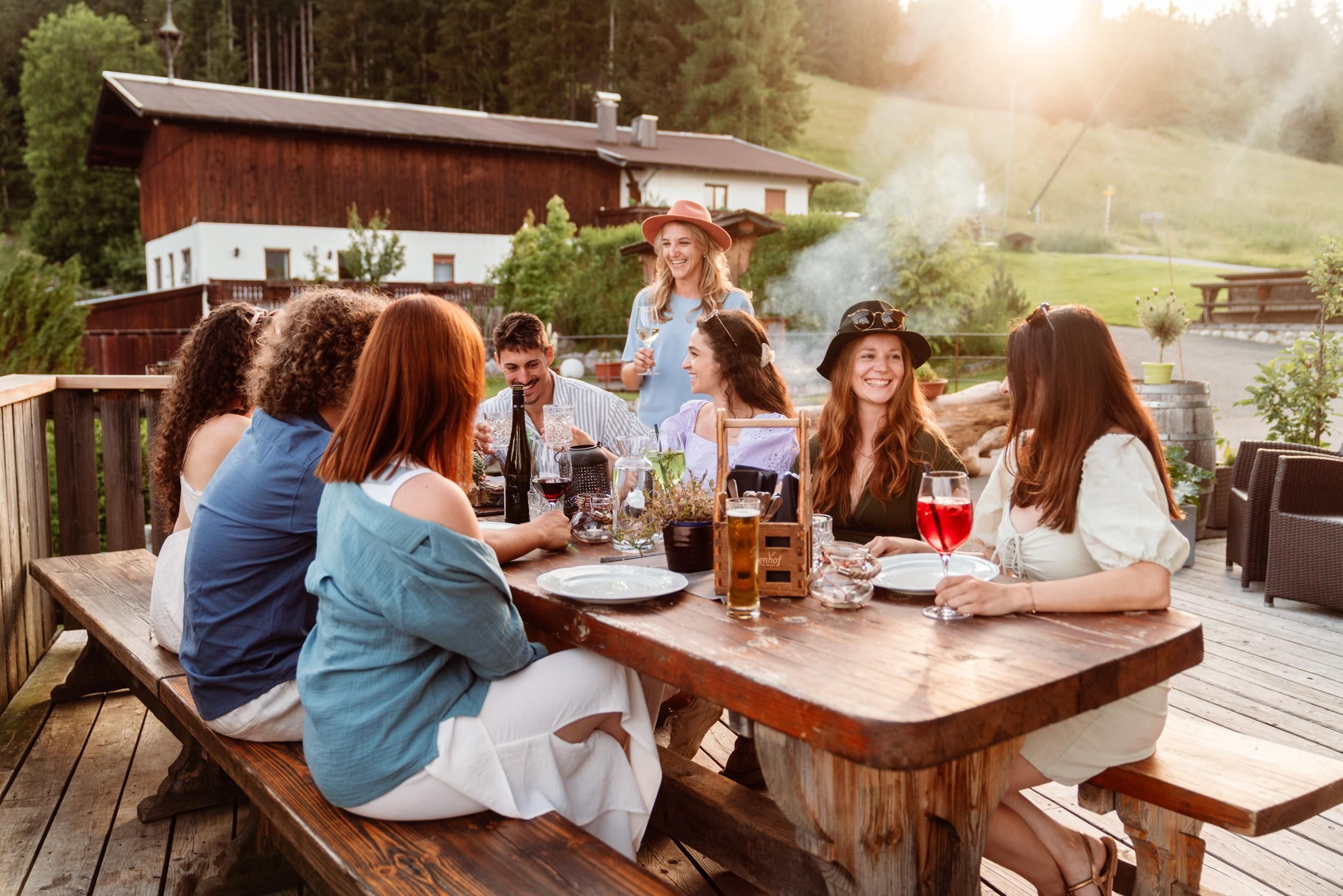 A group of friends is sitting at a wooden table outdoors, enjoying drinks. In the background, a beautiful house and nature can be seen.