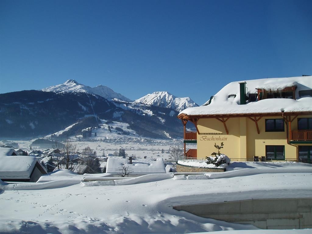 A picturesque winter landscape with snow-covered mountains and a charming building in the foreground. The clear blue sky complements the peaceful atmosphere.